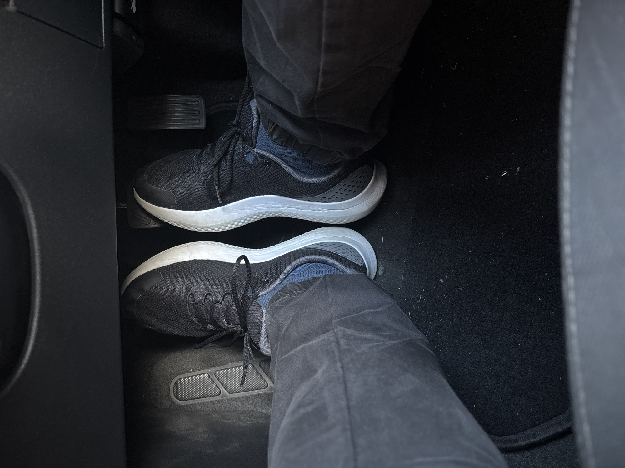 The image shows a first-person perspective of a person's feet wearing black sneakers inside a vehicle, likely a car. The feet are positioned on the floor of the car, with one foot resting on what appears to be a pedal. The pedals are dark, ribbed, and standard for a manual transmission vehicle. The sneakers are black with white soles and dark laces. The person is wearing dark-colored pants. The car's interior is dark and appears to be a standard driver's side footwell. There is no discernible information about the location within the city of Cork, Ireland, or any other geographical markers in the image. The scene is static and offers no indication of action or interaction beyond the placement of the feet.
