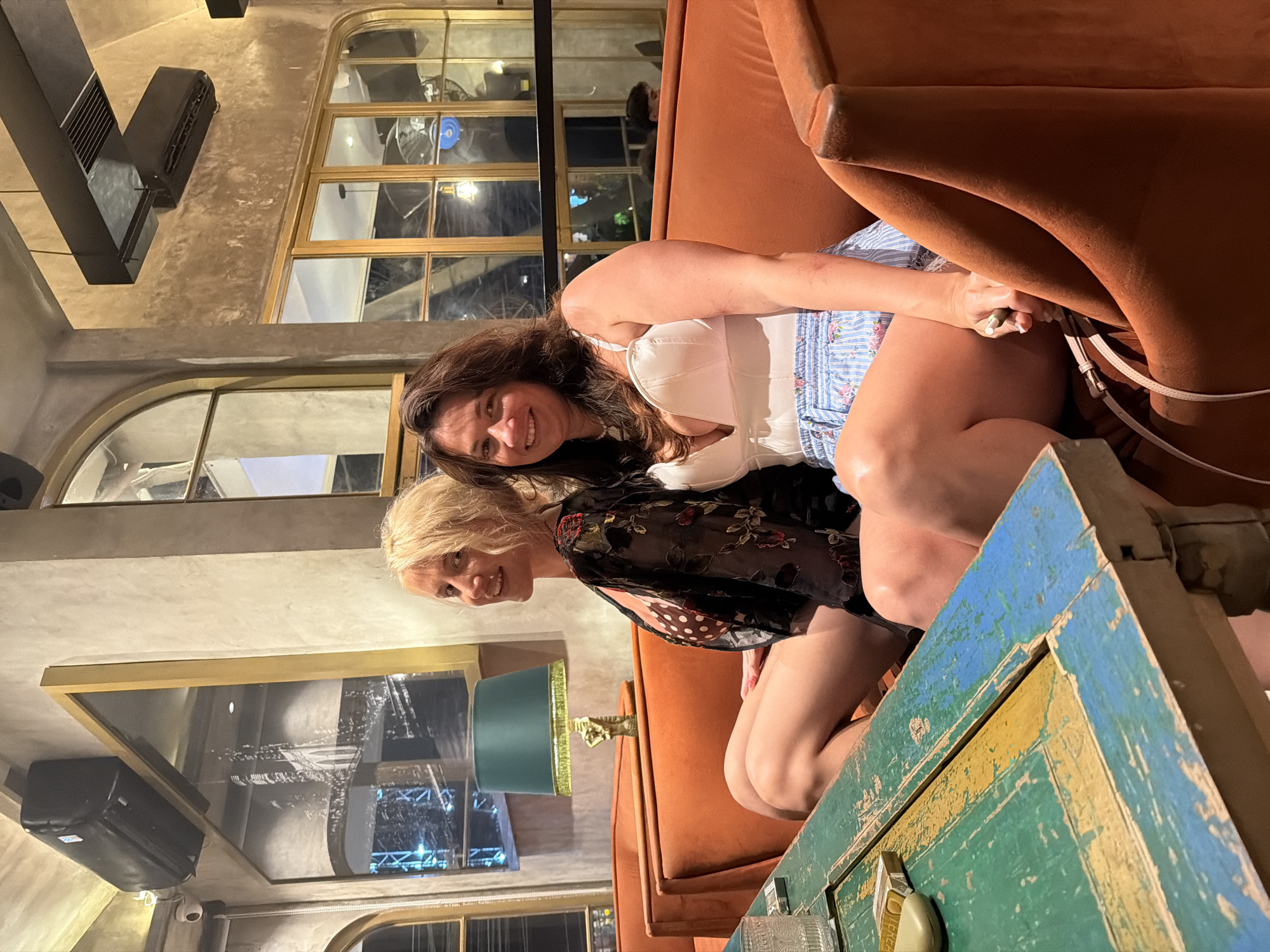 Two women are seated on an orange velvet couch in what appears to be a dimly lit, upscale restaurant or bar in North Kuta, Indonesia. The woman on the left has blonde hair and is wearing a black lace top with a polka dot bikini top underneath. The woman on the right has dark hair and is wearing a white corset-style top and blue and white striped shorts with a floral pattern. Both women are smiling and looking at the camera. In the foreground, a distressed, multi-colored wooden table with green, blue, and yellow paint is visible. The background features ornate mirrors, large arched windows, and decorative lighting, suggesting an ambient and possibly exotic atmosphere. An air conditioning unit is mounted on the wall above a large framed mirror.