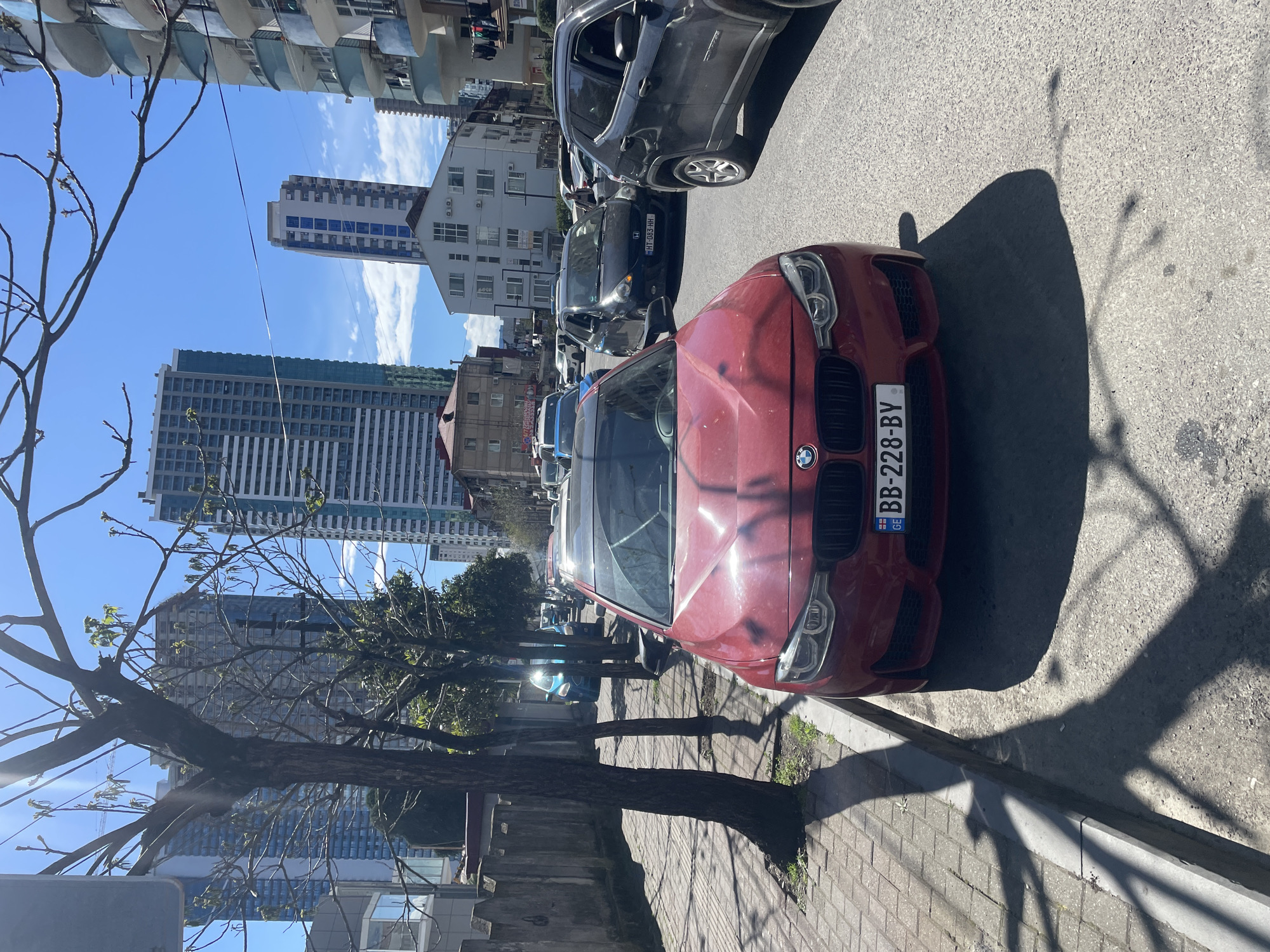The image shows a street scene in Batumi, Georgia. A red BMW is parked on the side of the road. Several other cars are parked in front of and behind the BMW. Tall buildings are visible in the background, and trees line the street. The sky is blue with some clouds. The weather is sunny. The street is paved, and there is a sidewalk. Shadows from the tree and the car are cast on the road.