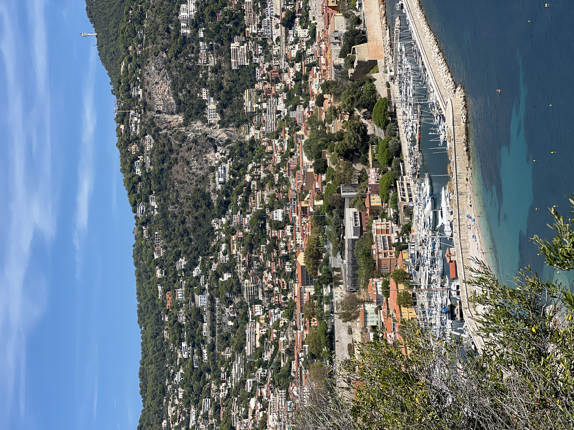 The image shows a panoramic view of the city of Nice, France, with a focus on its coastline and hillside architecture. In the foreground, lush green foliage partially obscures the view. Below, a vibrant blue sea is dotted with yellow buoys. A prominent stone pier juts out into the water, lined with numerous sailboats and yachts docked in a marina. A small sandy beach is visible along the inner edge of the pier, with a few people sunbathing.
The city itself sprawls up a steep, tree-covered hillside. Buildings with terracotta-tiled roofs are densely packed together, interspersed with green trees and patches of rocky terrain. Many of the houses are white or light-colored, with some featuring prominent balconies and windows. Higher up the hill, more dispersed villas and residential buildings are visible, blending into the natural landscape. A tall telecommunications tower stands on the peak of the furthest hill, overlooking the entire scene. The sky is a clear, bright blue with a few wispy white clouds.