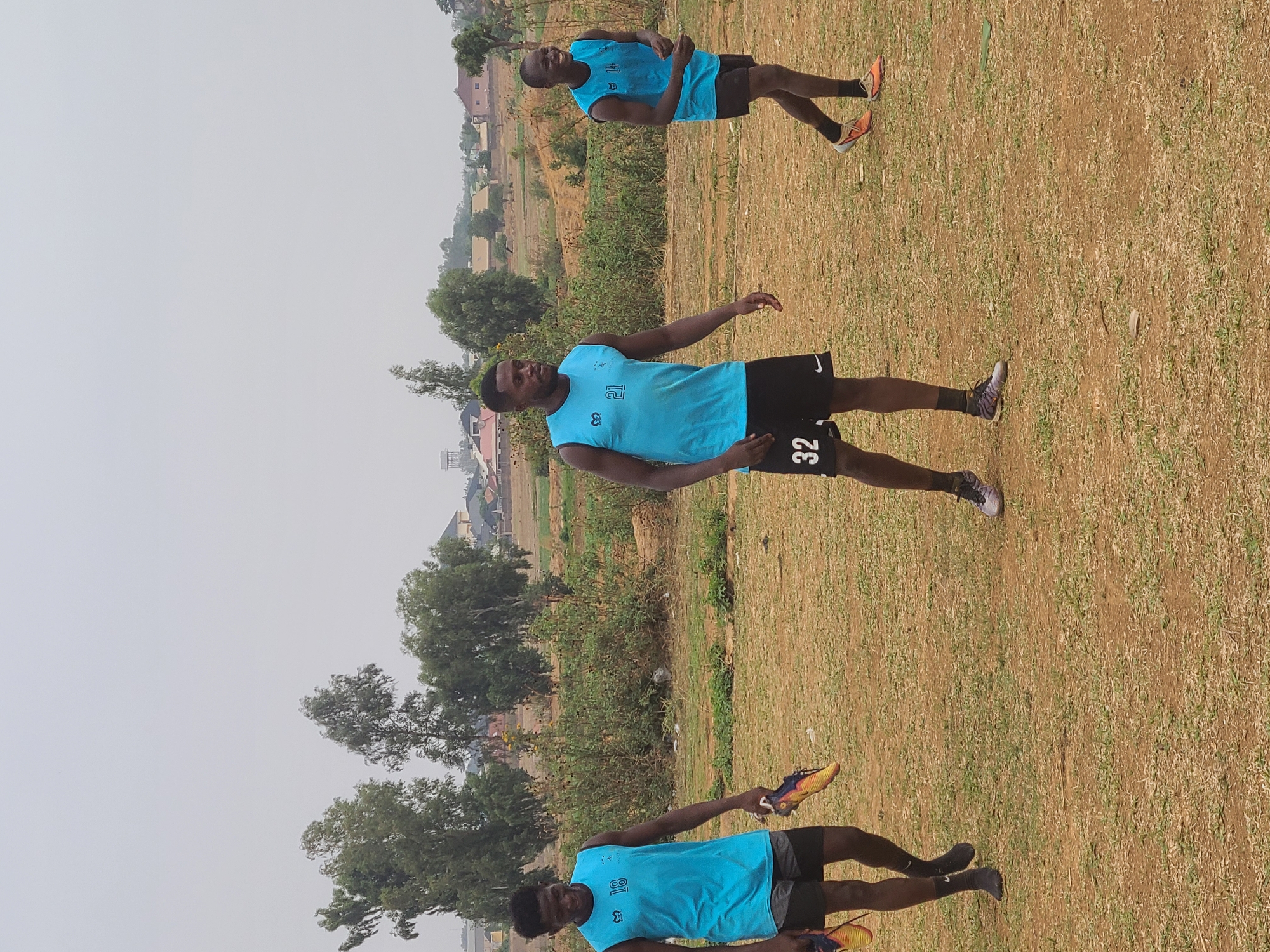 The image shows three men in a field in Barikin Jirga Summa, Nigeria. All three men are wearing light blue athletic jerseys and black shorts. The man in the foreground is holding a pair of running shoes in his right hand. The man in the middle is standing with his arms outstretched. The third man is further in the background. The field is brown with patches of green grass. Trees and buildings are visible in the distance. The sky is overcast.