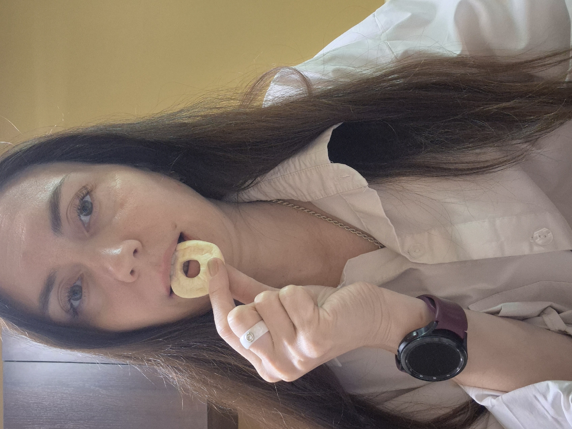 A woman is shown in a close-up, slightly angled shot. She holds a round, hole-centered object, possibly food, to her mouth. She wears a white collared shirt, a silver chain necklace, a white ring with a stone, and a black smartwatch with a purple strap. The background is a muted yellow wall and a dark brown surface. The location is Unknown, Unknown.