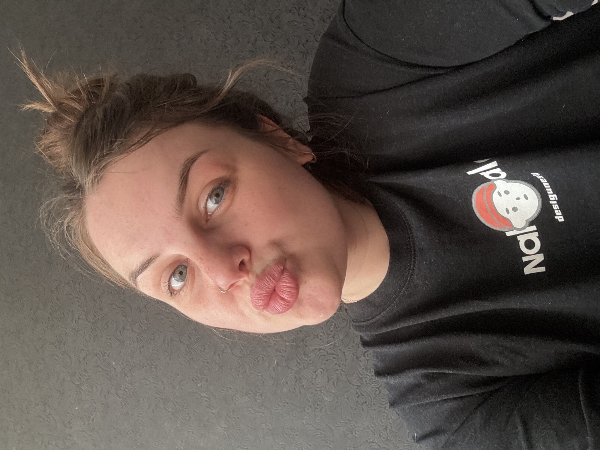 A woman with light skin, light eyes, and a nose ring is positioned facing the camera with her lips pursed in a kiss. Her hair is gathered in a messy bun. She wears a dark-colored t-shirt with white lettering and a graphic on the chest. The background is a dark, textured surface. No location in the city of Chornomorskoe, Ukraine is detectable.