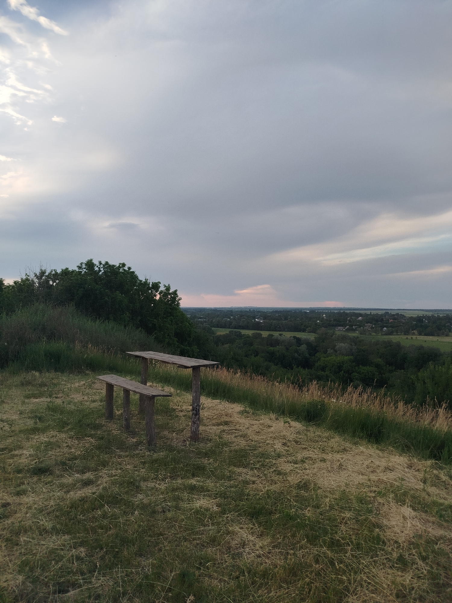 This is a medium-shot, outdoor, eye-level, static photograph taken from a grassy hilltop overlooking a rural landscape. In the foreground, a rustic wooden bench and a matching picnic table are positioned on a patch of dry, yellowed grass interspersed with green vegetation. The bench is simple, made of rough-hewn planks supported by three thick wooden legs. The picnic table shares a similar construction, with a long plank surface supported by two sturdy wooden posts. Behind the seating area, a dense line of dark green trees forms a natural barrier, leading to a broader vista. The midground displays a hazy expanse of rolling hills covered in a patchwork of green trees and scattered buildings, suggesting a village or small town. The horizon is softened by a light atmospheric haze. The sky above is overcast with a dramatic gradient of gray and white clouds, with a subtle hint of pink light breaking through on the left side, indicating either sunrise or sunset. The overall impression is one of a quiet, natural resting spot with a scenic view in a rural area. The image is located in the village of Ustyvytsia, Ukraine.