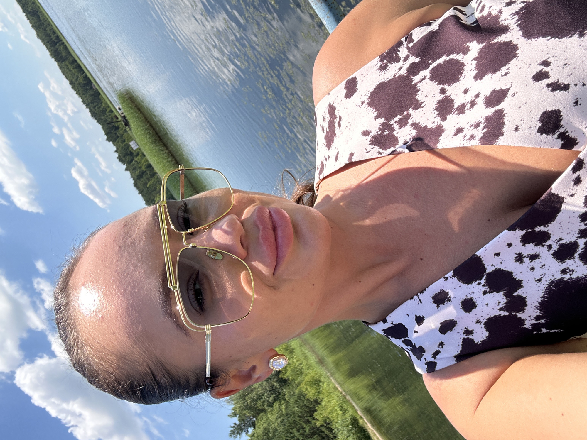 In a selfie taken outdoors, a woman with her hair pulled back and wearing large, gold-rimmed sunglasses smiles at the camera. She is wearing a black and white patterned top. A lake is visible in the background, with trees lining the far shore. The sky is blue with scattered white clouds. The location is the city of Duvankul', Russia.