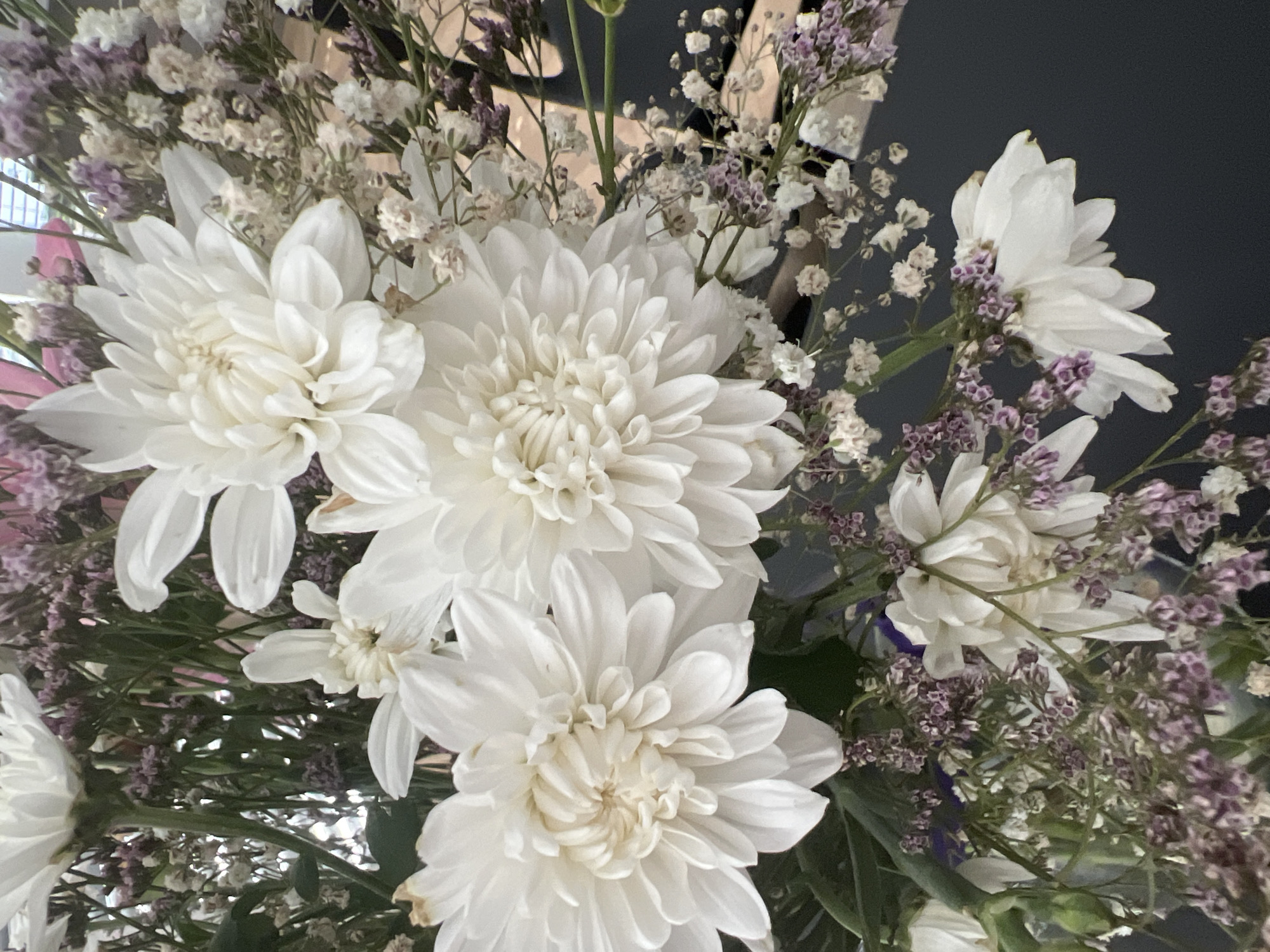 A close-up, overhead shot shows a bouquet of white dahlias and smaller purple and white filler flowers. The bouquet is arranged in a dark grey vase, which is not fully visible. The background is dark and blurred. There are no people in the image. The image is focused on the floral arrangement, highlighting the textures and colors of the flowers. The location of Hof HaCarmel Regional Council, Israel is not depicted or implied in the image.