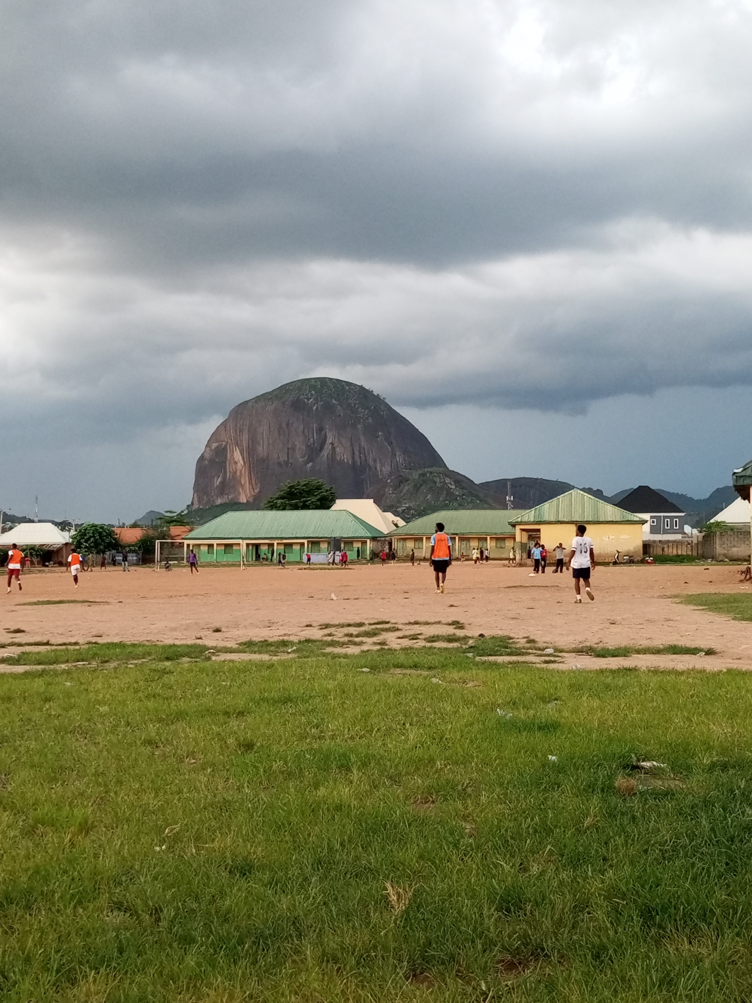 In Sabojibi, Nigeria, an outdoor soccer game is in progress on a dirt field. The field is bordered by a grassy area in the foreground and a line of green-roofed buildings in the background. In the distance, a large, rocky mountain looms under a dramatic, cloudy sky. Several individuals, mostly young men and boys, are visible on the field, some engaged in the game and others observing. The players are dressed in athletic attire, including shorts and t-shirts, with a few wearing bibs to distinguish teams. The overall atmosphere appears to be that of a casual community sporting event.