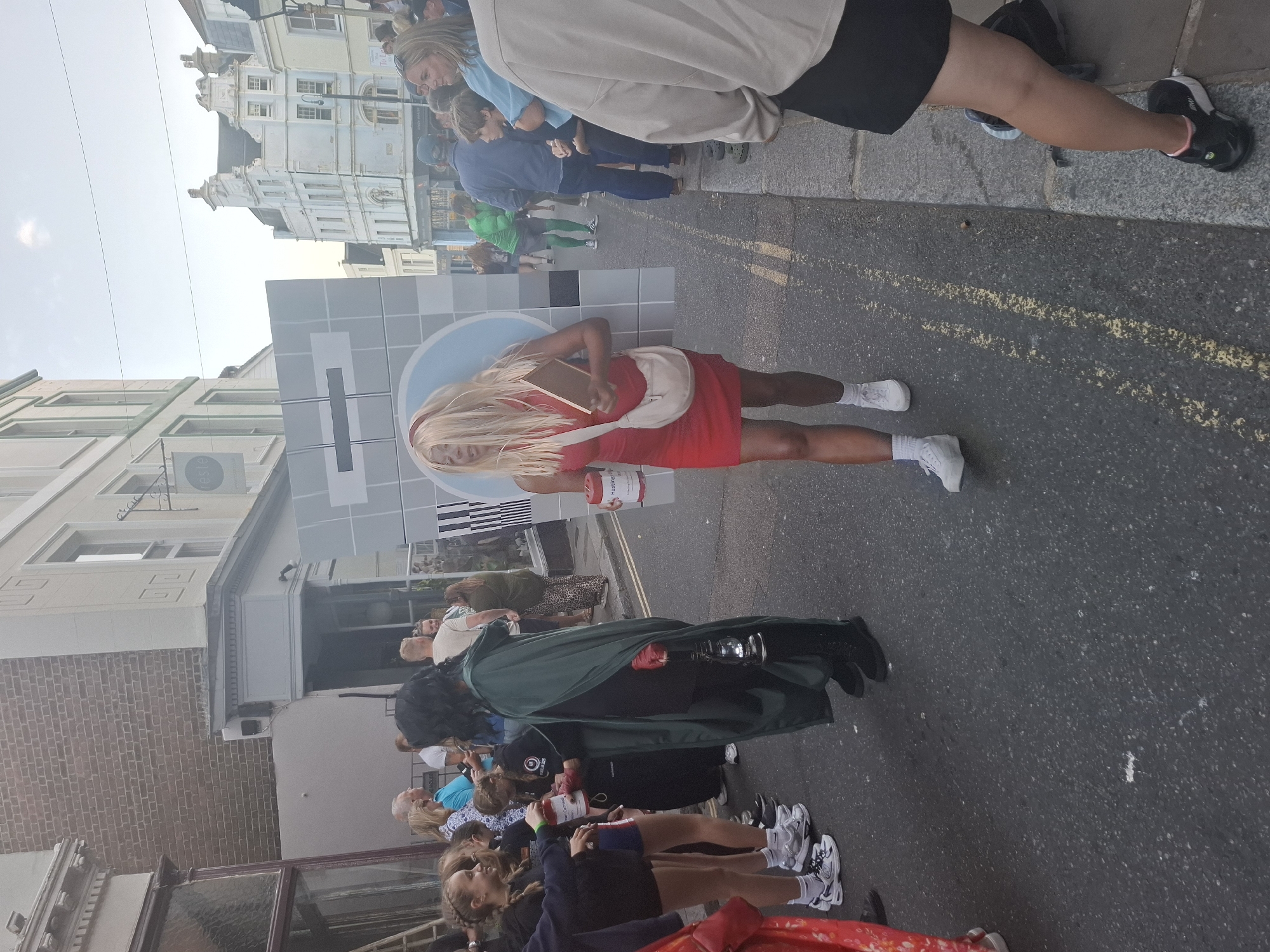 In St Leonards, United Kingdom, a person in a red dress and blonde wig, carrying a large cardboard television test pattern, walks down a street. They are holding a red cup and a book. A person in a green cape and black boots, holding a lantern, walks beside them. Other people, dressed in casual clothing, are visible in the background, some of whom appear to be watching the procession. The scene is set on a street with buildings on either side and a gray sidewalk. The time of day appears to be late afternoon or early evening, given the lighting conditions.