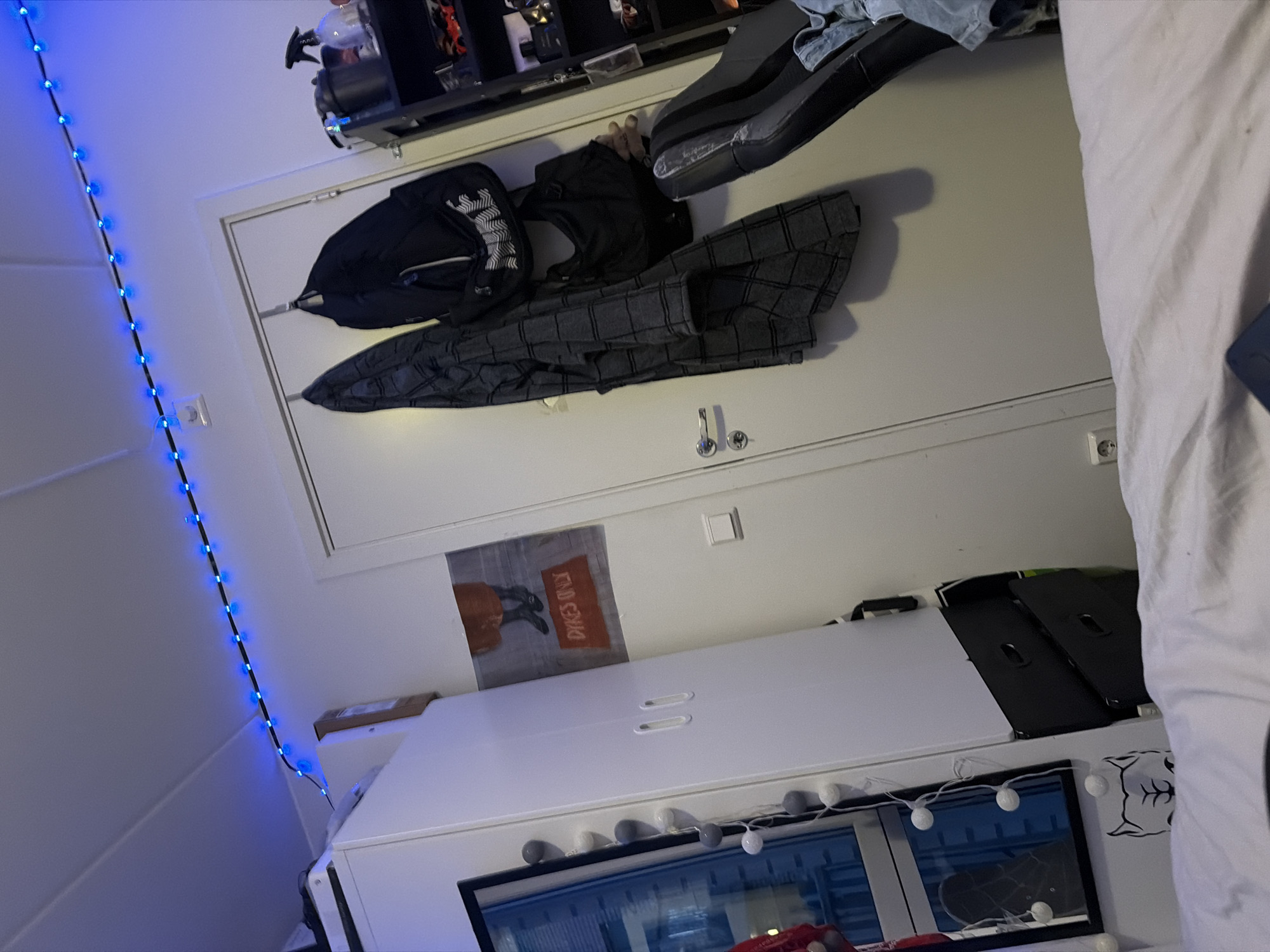 This is a medium shot, eye-level, indoor photograph taken in what appears to be a bedroom. The scene is dominated by a white door with a silver doorknob. On the door, there are two items hanging: a dark patterned jacket or robe on the left and a black backpack on the right. Below the backpack, a black duffel bag is also hanging.
To the left of the door, a large white wardrobe with two handles is visible. A poster with a person's legs and the text "DYKES ONLY" is affixed to the wall between the wardrobe and the door. String lights with small, round white and grey bulbs hang vertically down the side of the wardrobe. A framed picture is visible within the wardrobe's reflection.
To the right of the door, a black shelving unit is visible, holding various objects including what appears to be spray bottles and containers. A black office chair with a distressed seat is positioned in front of the shelving unit. The background is a pale-colored ceiling with blue LED strip lights running along the edge. The foreground shows a white duvet or comforter. The overall impression is of a lived-in, casual space.
There is no information within the image that indicates the specific location of Oslo, Norway.