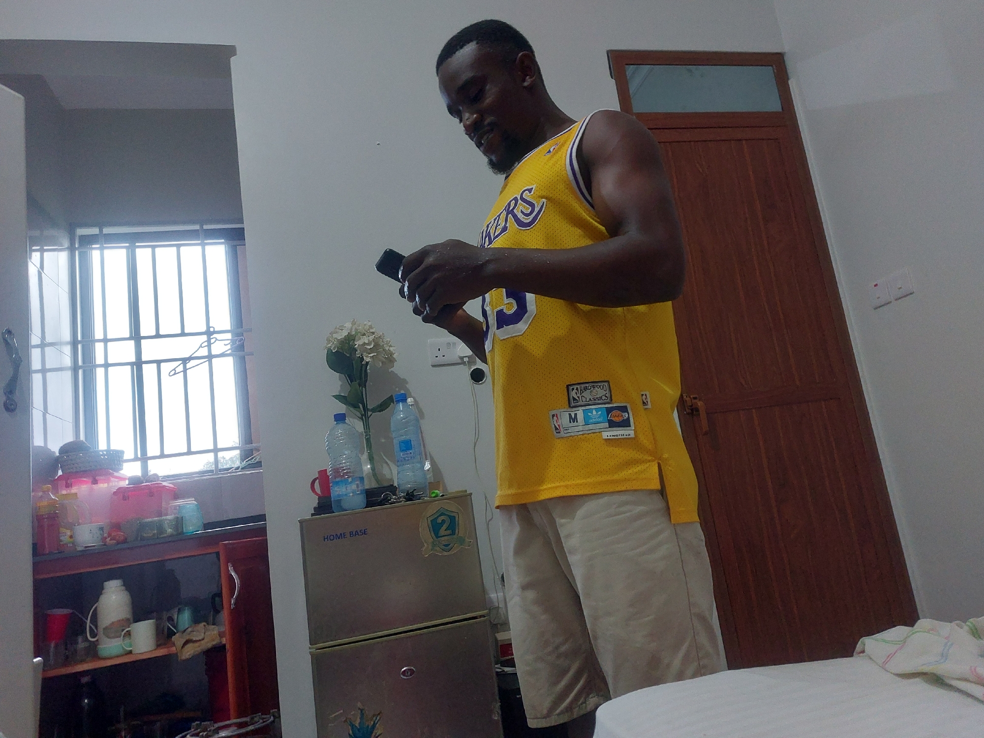 A man in a yellow Lakers jersey and beige shorts is standing in a room. He is looking down at a black smartphone in his hands. Behind him is a dark brown wooden door with a windowpane at the top. To the left, a doorway leads to a room with a barred window, a shelf filled with various containers, and a mini-fridge labeled "HOME BASE." There are water bottles and a vase of flowers on top of the mini-fridge. A bed is partially visible in the foreground on the right. The environment appears to be a modest residential interior. There is no information that can confirm the location in the city of Dar es-Salaam, Tanzania.