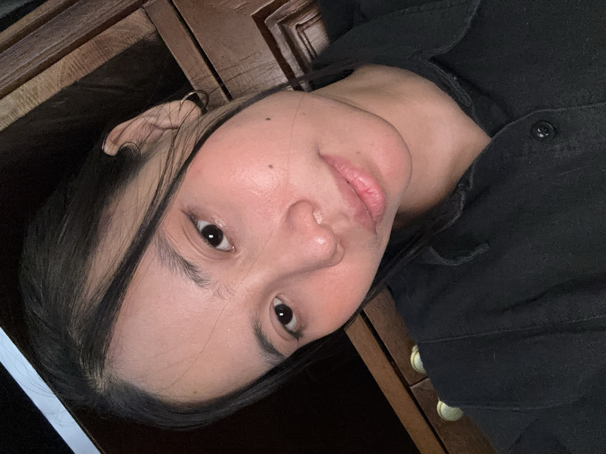 A close-up, high-angle selfie of a young East Asian woman with dark hair, dark eyes, and light skin. She is wearing a black collared shirt. Her hair is pulled back, with some strands falling around her face. She has a faint blush on her cheeks and a small beauty mark on her right cheek. The background appears to be a wooden surface, possibly furniture. The lighting is soft and casts a slight glow on her face. There is no discernible urban environment or specific location within South Jakarta, Indonesia.