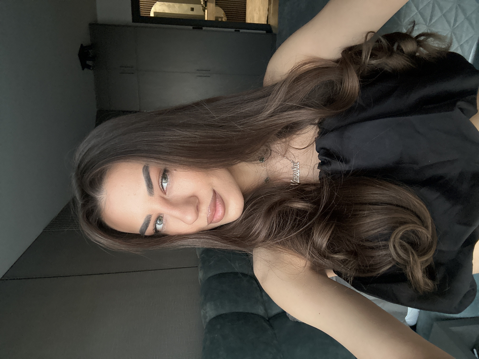 A woman is taking a selfie in what appears to be a modern apartment bedroom in Sofia, Bulgaria. She has long, dark brown, wavy hair and is wearing a black, strapless top. A silver necklace with a nameplate is visible around her neck. She is looking directly at the camera with a neutral expression. In the background, there is a large, dark upholstered headboard and a wardrobe. The lighting is soft, suggesting it may be daytime or evening.