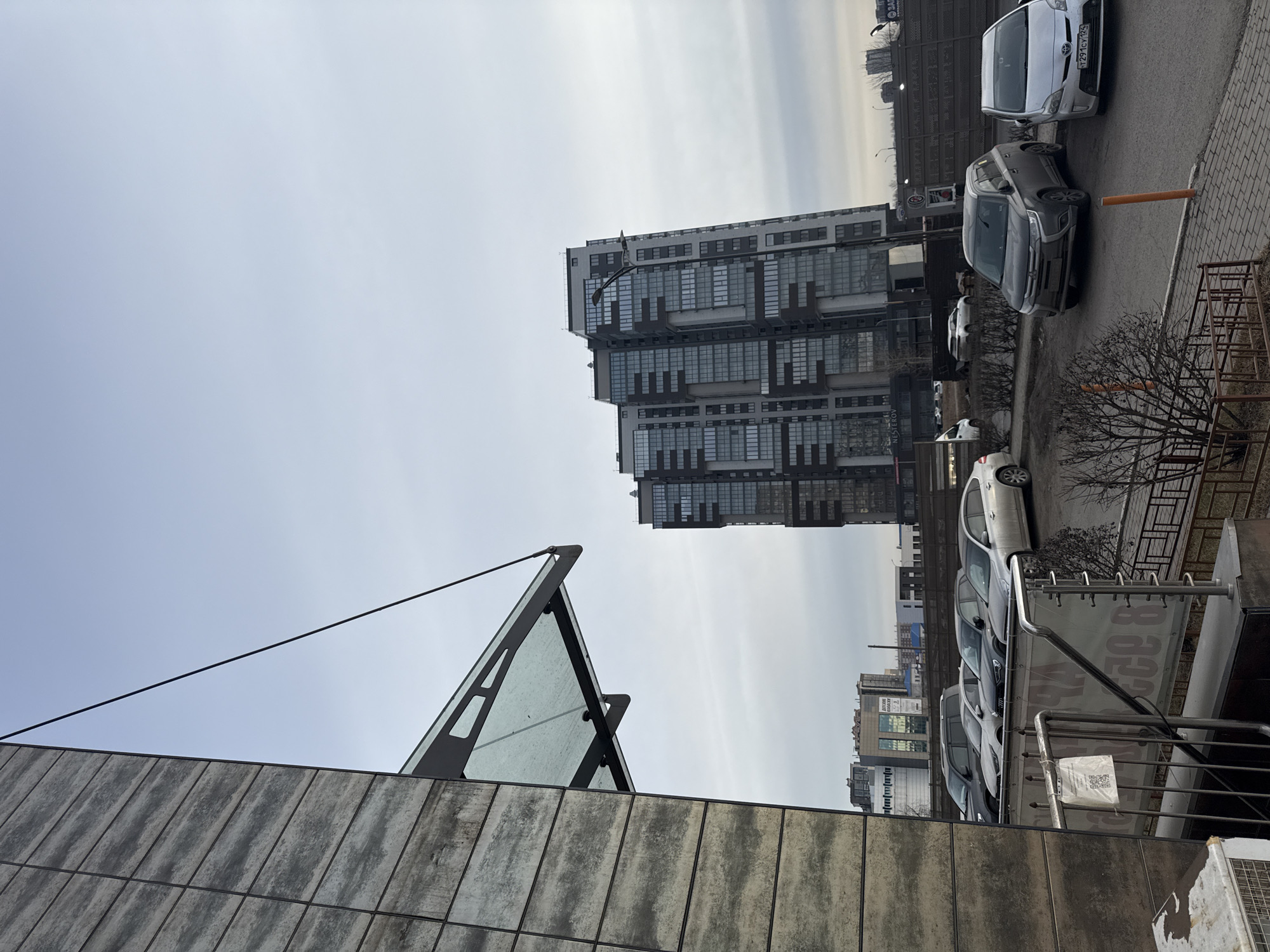 The image shows an urban scene in Krasnoyarsk, Russia, featuring a mix of architectural styles and vehicles. In the foreground, there's a modern building with glass and concrete elements. Above this, a tall, multi-story building with a grid-like facade dominates the skyline. To the right, a row of parked cars lines the street, and a metal railing is visible in the foreground. The sky is overcast with a hint of sunlight breaking through. Overall, the scene depicts a typical urban environment with a blend of modern and older structures.