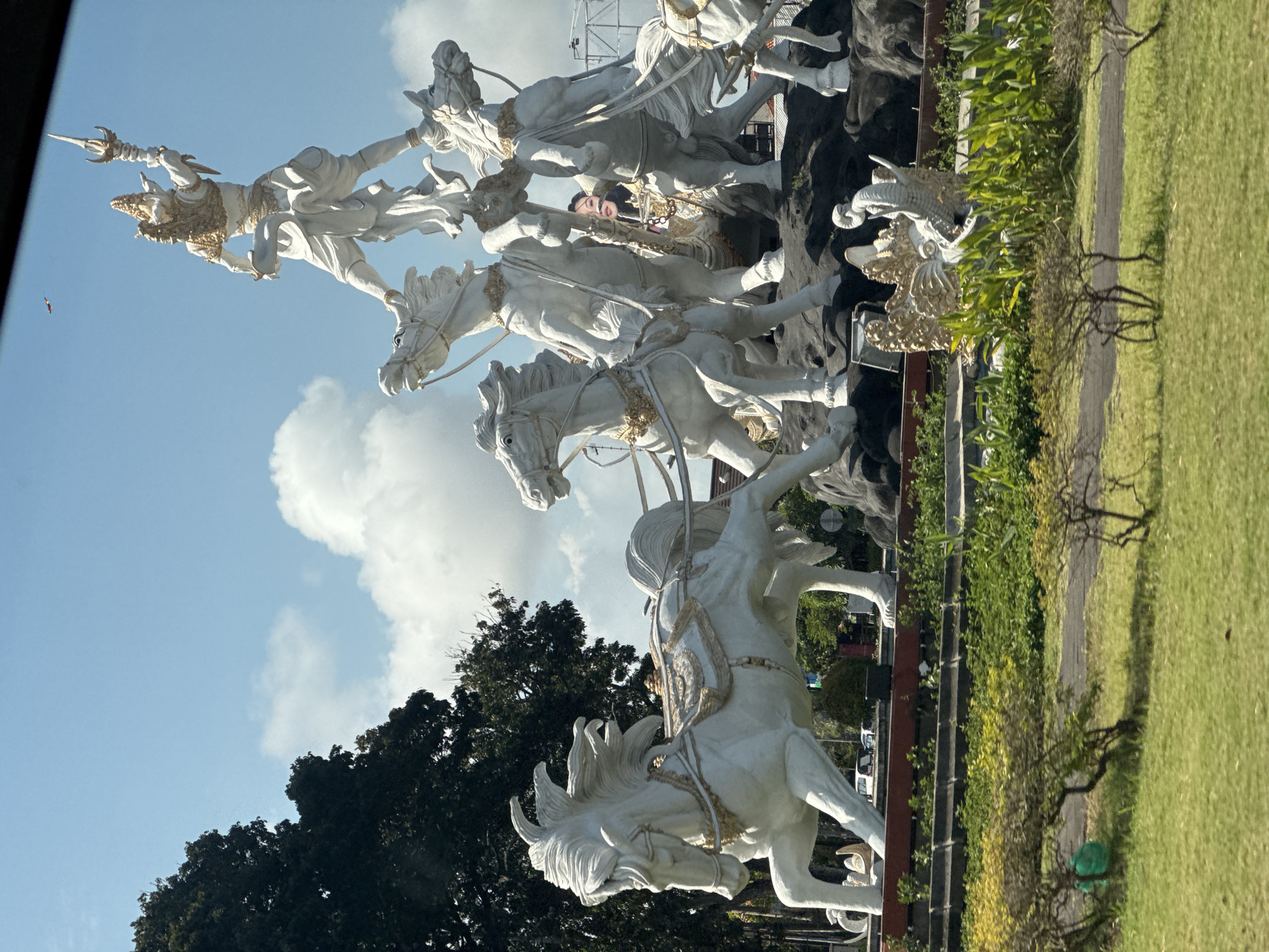 This image captures the iconic Catur Mukha statue located in Kuta, Bali, Indonesia. The statue depicts a deity, likely from Balinese mythology, standing atop a chariot pulled by several white horses. The deity is rendered in a dynamic pose, holding a weapon aloft. The horses are also detailed, with flowing manes and ornate harnesses. In the background, lush green trees and a bright blue sky with scattered clouds form the natural environment. The foreground shows a well-maintained grassy area with some landscaping. A person is visible in the statue, looking out from behind the horses. The overall impression is one of grandeur and cultural significance, set against a tropical backdrop.