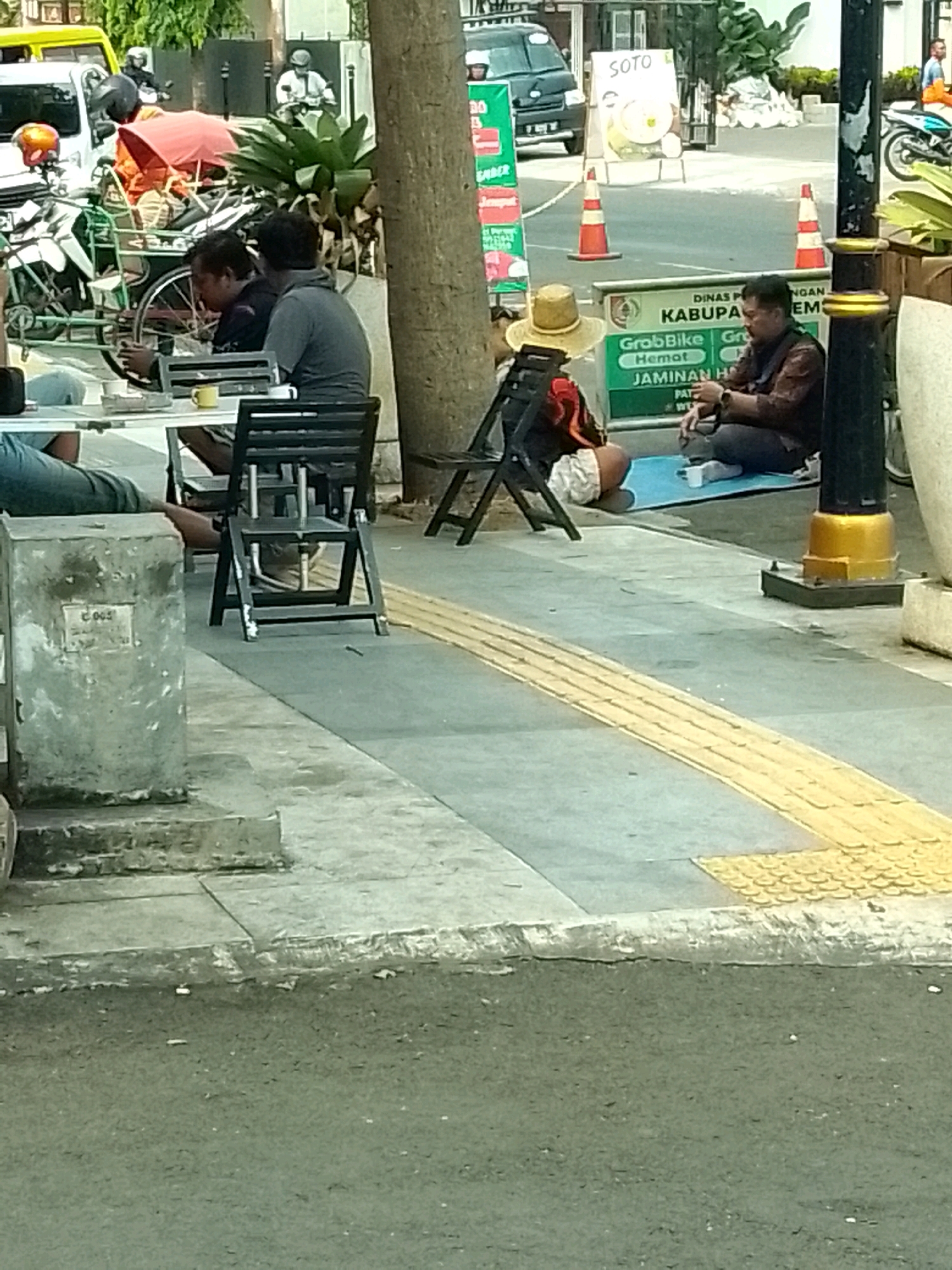 This is a street-level outdoor photograph taken in Jember, Indonesia. The image depicts a sidewalk scene with several individuals and various objects. In the foreground, a light gray concrete structure with a plaque is visible. Behind it, a white table with a yellow cup and what appears to be a cigarette butt are present. A black folding chair is positioned to the right of the table.
Further back, on the sidewalk, a person wearing a straw hat and a red and black shirt is seated, facing away from the camera. To their right, a person in a brown shirt and dark pants is seated on a blue mat, holding a cigarette. A black folding chair is behind the person in the straw hat.
In the background, a large tree trunk stands to the left of the seated individuals. Beyond the sidewalk, a street is visible with traffic cones, a "SOTO" sign on a board, and a dark-colored van. To the far right, a motorcycle with a rider is parked near a lamp post with gold accents. Another motorcycle is visible on the far right edge of the frame. The ground is paved with gray concrete and features a raised yellow tactile paving for the visually impaired.