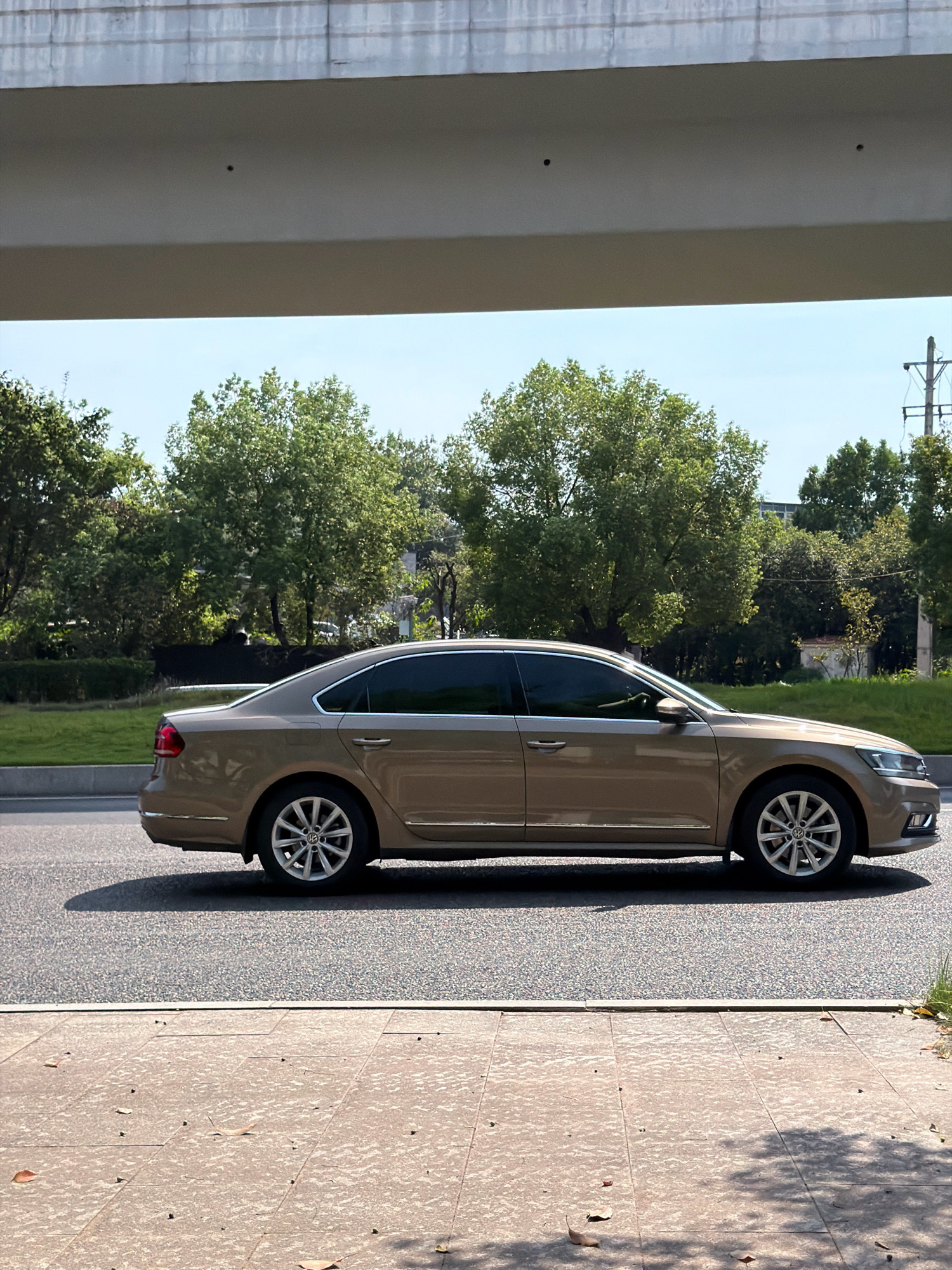 A tan sedan is parked on the side of a road in Jiangjin, China. The car is facing right, and its windows are tinted. The road is paved with asphalt, and there is a sidewalk on the right side of the road. In the background, there are trees and a blue sky. The scene is illuminated by natural sunlight, casting shadows on the road and sidewalk. The overall environment appears to be an urban or suburban setting, possibly on a sunny day.