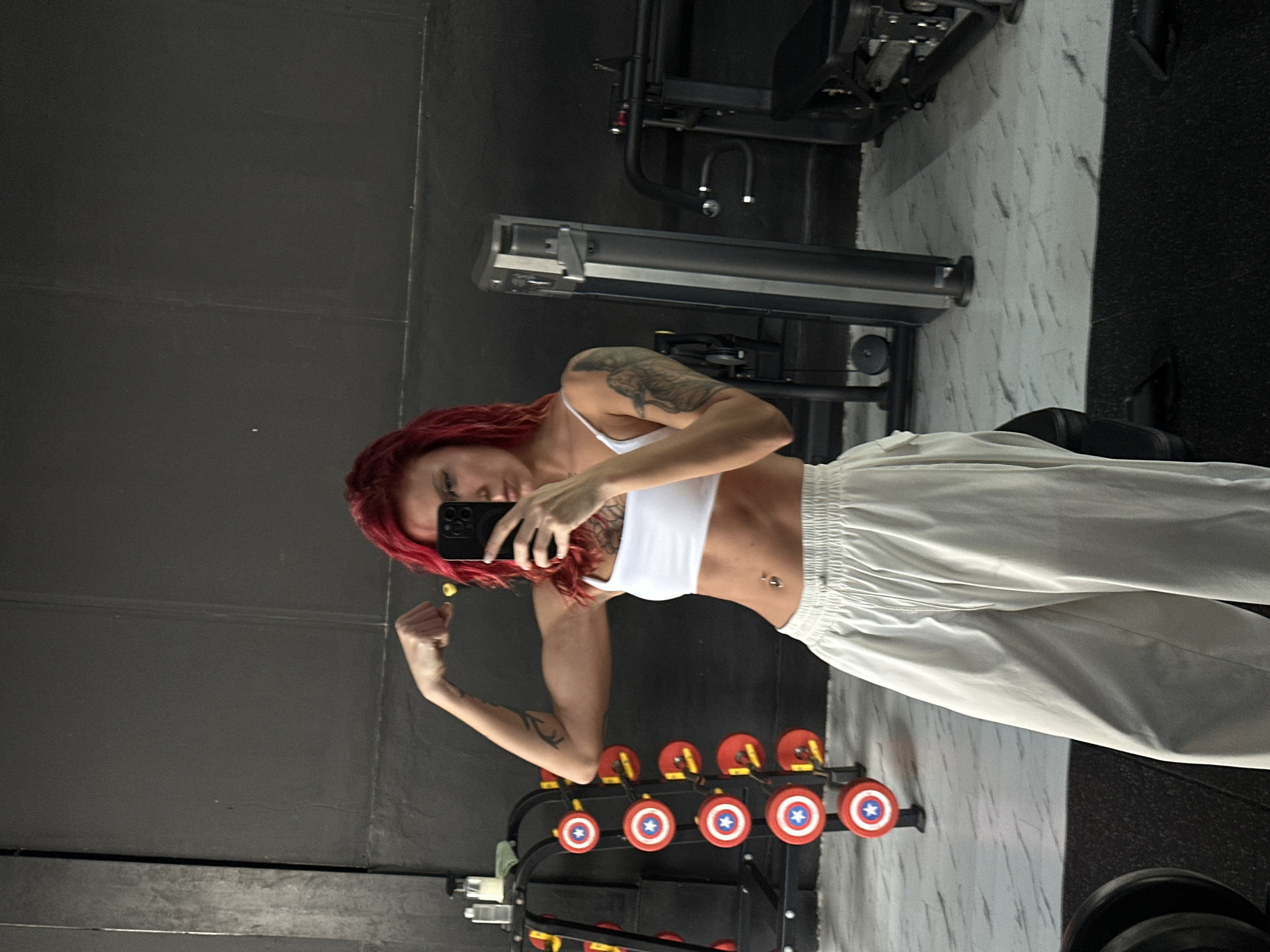 A woman with dyed red hair and tattoos is taking a selfie in a gym in North Kuta, Indonesia. She is wearing a white sports bra and white pants. She is standing in front of a weight rack with weights resembling Captain America shields. There is gym equipment visible in the background, and the floor appears to be tiled. The lighting is bright.