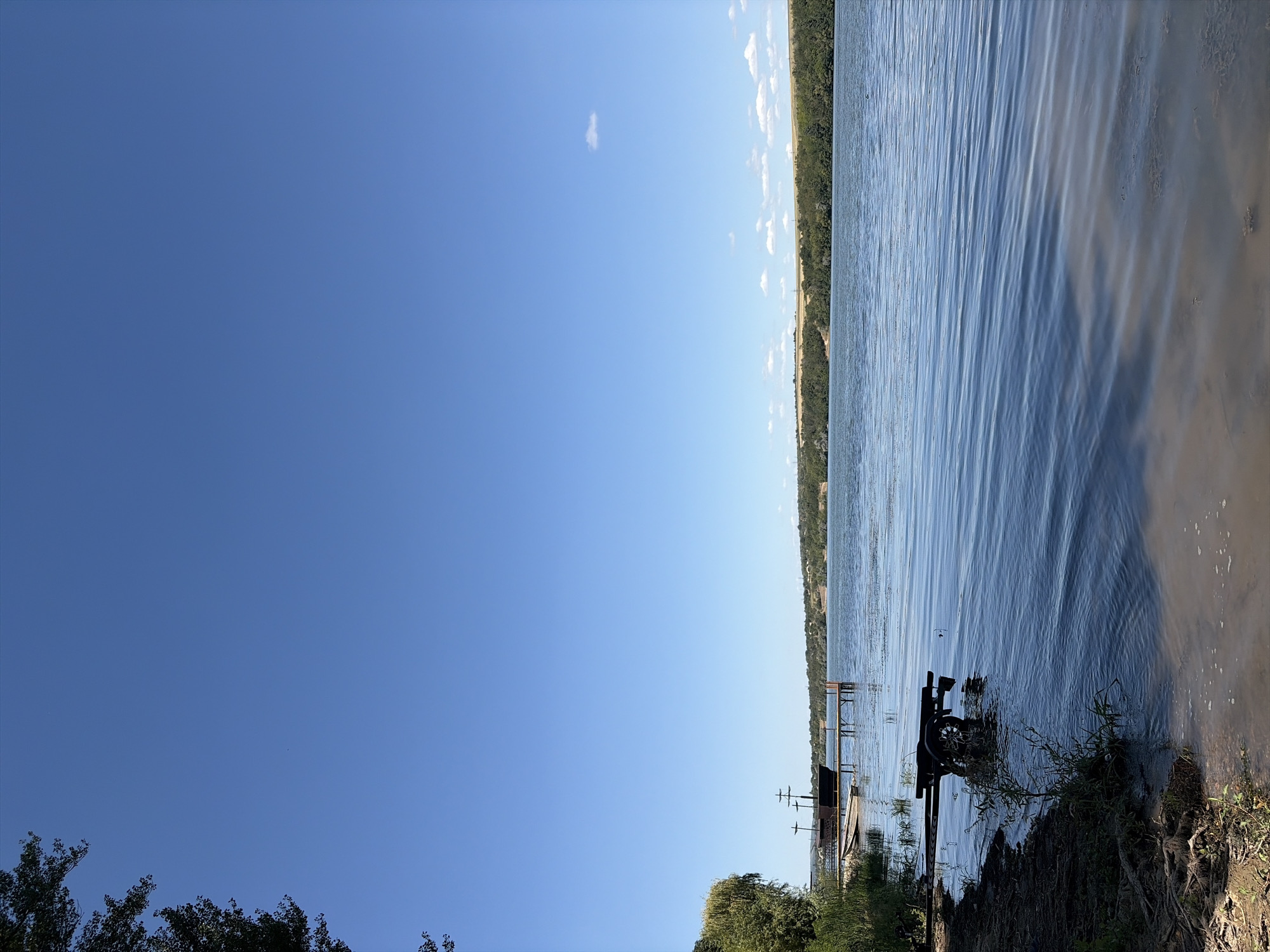 The image depicts a serene waterscape, likely a lake or river, under a clear blue sky with a few wispy clouds. The foreground features a sandy and grassy shoreline with visible tree roots. Partially submerged in the shallow water near the bank is a dark-colored boat trailer. To the left, lush green vegetation and the branches of a large tree extend into the frame. In the mid-ground, a wooden pier juts out into the water, and a decorative boat, resembling a pirate ship, is moored alongside it, accompanied by a small rowboat. The opposite bank is lined with dense green trees and rolling hills, with hints of structures and vegetation in the distance. The water surface is calm with gentle ripples reflecting the sky. The overall impression is one of a tranquil natural setting.
