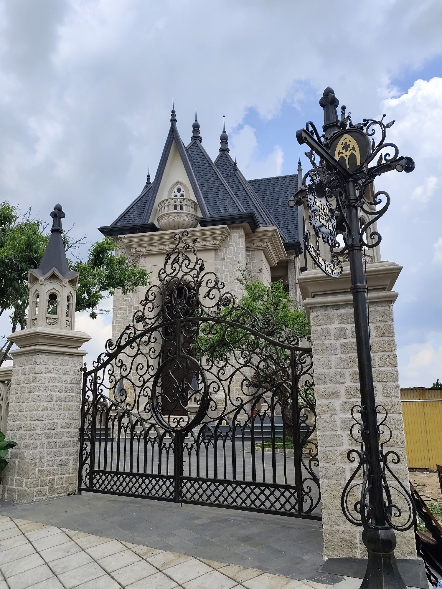 The image features a grand, ornate metal gate leading to a building designed in a European architectural style, resembling a small castle or mansion. The building is characterized by gray stone walls, a dark gray, steeply pitched roof with multiple turrets, and cream-colored trim. An attached building with a turret and black steeple sits to the side. The metal gate is intricately designed with swirling patterns and vertical bars, flanked by stone pillars. A black metal lamppost with decorative scrolls and a sign with the words "ROMAN CASTLE 22/22 Le Hong Phong Da Lat City" stands to the right of the gate. The scene is set in Da Lat, Vietnam, as indicated by the text on the sign. The foreground consists of a paved walkway with gray and white bricks. The sky is overcast with clouds. The gate is closed, and there are trees partially visible behind the gate and the building. A yellow building can be partially seen on the right-hand side of the image.
