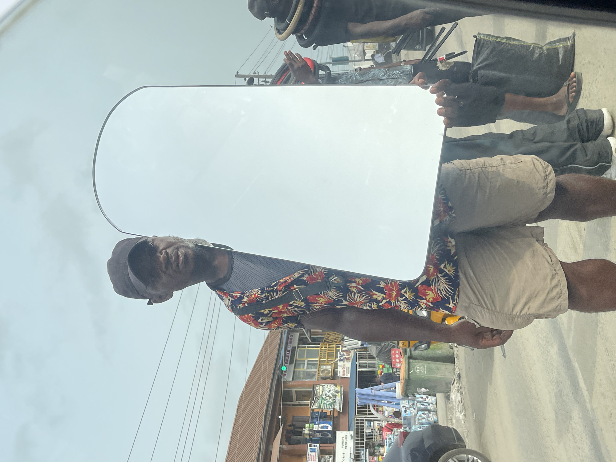 This media file captures a vibrant daytime street scene in Ado, Nigeria, viewed from the interior of a vehicle, likely through a window exhibiting reflections.
The central figure is a dark-skinned man, positioned prominently, wearing a black cap, a colorful floral short-sleeved shirt (blue with red and yellow patterns), and light-colored shorts. He holds a large, rectangular, plain white board or panel vertically with both hands, facing slightly towards the camera.
Behind him and the white board, a bustling urban street stretches out. Buildings with multiple shopfronts line the road. Visible signage includes "WORLD LIGHTS" on a green sign and "WURKUS VENTURES" on a blue sign. Green waste bins are placed along the roadside, and numerous power lines are strung across the sky. Several other people are visible in the background, engaged in daily street activities, appearing as vendors or passersby. Portions of other vehicles, including the rear of a dark car, are also visible.
A notable detail is the presence of reflections on the vehicle's window, particularly in the upper part of the image. These reflections show inverted figures of people, including one person holding a large, circular, wooden object, and another in dark attire. These reflections add a layer of visual complexity, indicating the photograph was taken through a reflective surface. The bright, possibly overcast sky suggests it's a clear day. The scene depicts a lively, everyday street environment.