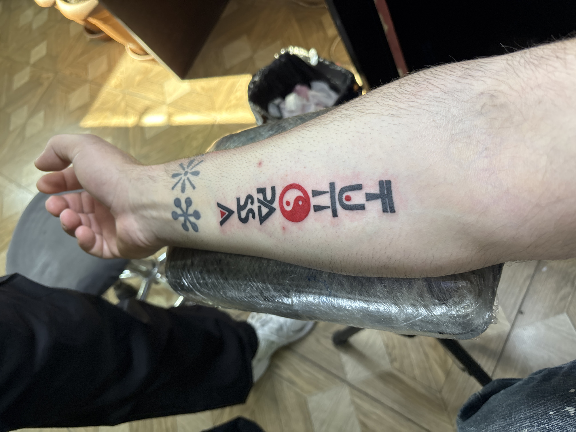 A person's forearm rests on a surface covered with plastic wrap. Visible on the forearm is a vertical series of tattoos, starting near the wrist with a design resembling stylized snowflakes or asterisks, followed by geometric shapes possibly representing letters or symbols, a red yin-yang symbol, and a final set of geometric shapes. The person's hand is also visible. The person is wearing black pants and white sneakers. A metallic trash bin is in the background. The scene is lit by artificial light, suggesting an indoor setting. There is no information to infer that this image was taken in Tbilisi, Georgia.