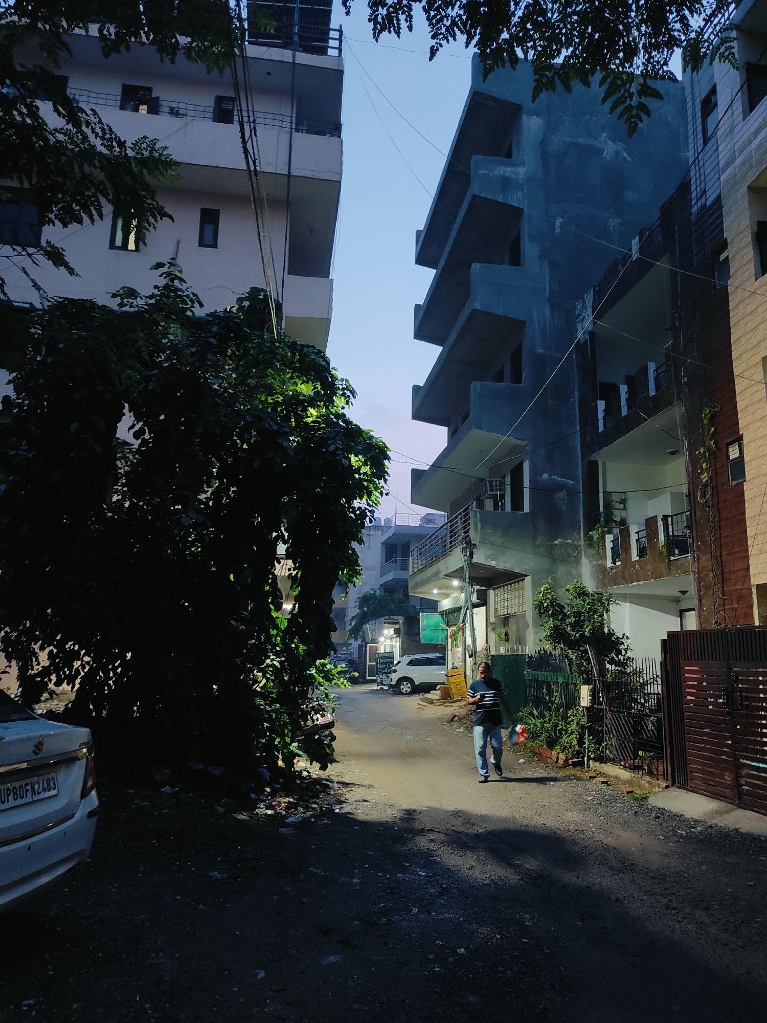 The image depicts a street scene in Gurgaon, India, likely during the early evening or late afternoon given the lighting conditions. On the left, a tall, light-colored building with multiple balconies is partially obscured by lush green foliage. The building has several windows, some illuminated. Adjacent to it, on the right, is a darker, more modern building with cantilevered balconies and a rough, textured facade, also showing some signs of interior lighting.
The street itself is unpaved, covered in gravel and debris. A white sedan is parked on the left side of the street, with its license plate visible as "UP80FK2483". A single man is walking down the street towards the viewer, dressed in a striped shirt and light-colored pants. He is carrying a colorful pinwheel. Further down the street, more buildings and fences are visible, along with another parked vehicle and some shop signs. The sky above is a clear, pale blue, indicating a transition from daylight. The overall impression is one of a residential or mixed-use urban environment in India, with a focus on the architecture and the everyday movement of people and vehicles.