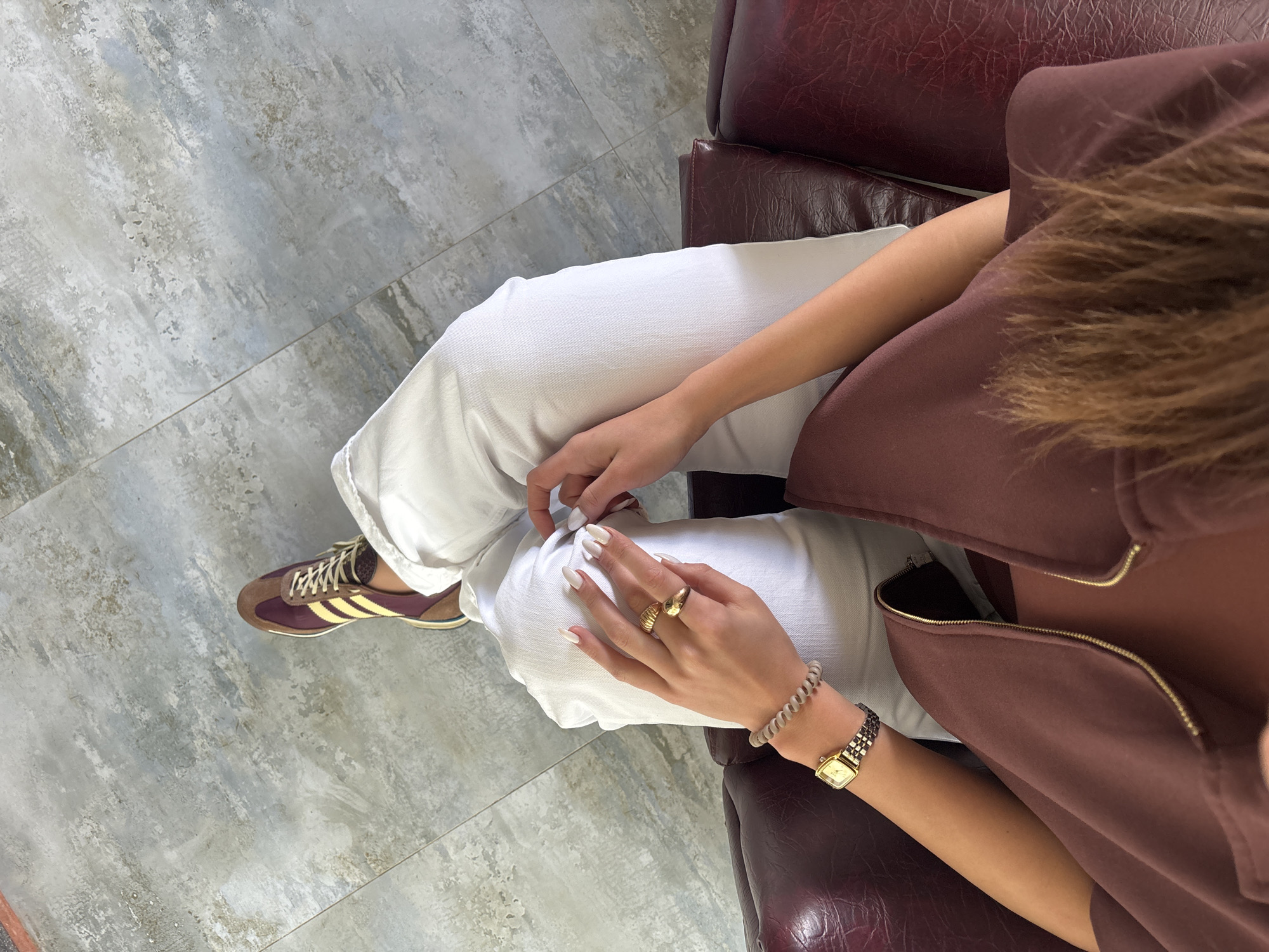 The image provides an overhead, slightly angled view of a person seated indoors, likely in a waiting area or lounge in Yerevan, Armenia. The flooring consists of large, light grey and off-white patterned tiles, possibly mimicking concrete or stone. The person is sitting on a dark reddish-brown (maroon) leather armchair or sofa, partially visible on the right.
The individual is wearing white, loose-fitting pants and a deep reddish-brown top or jacket with a golden zipper partially undone. On their left foot, a maroon sneaker with three yellow stripes (suggesting Adidas branding) and beige laces is visible. Their hands, adorned with long, white-painted fingernails, are lightly clasped on their lap. On the left hand, two gold-colored rings are worn: one on the ring finger and a larger, dome-shaped ring on the middle finger. The right wrist features a gold-colored watch with a square face and a metallic band, along with a light grey spiral hair tie. A section of light reddish-brown hair is visible over their right shoulder. The person appears to be in a relaxed posture, possibly resting or waiting. There is no visible text in the image.