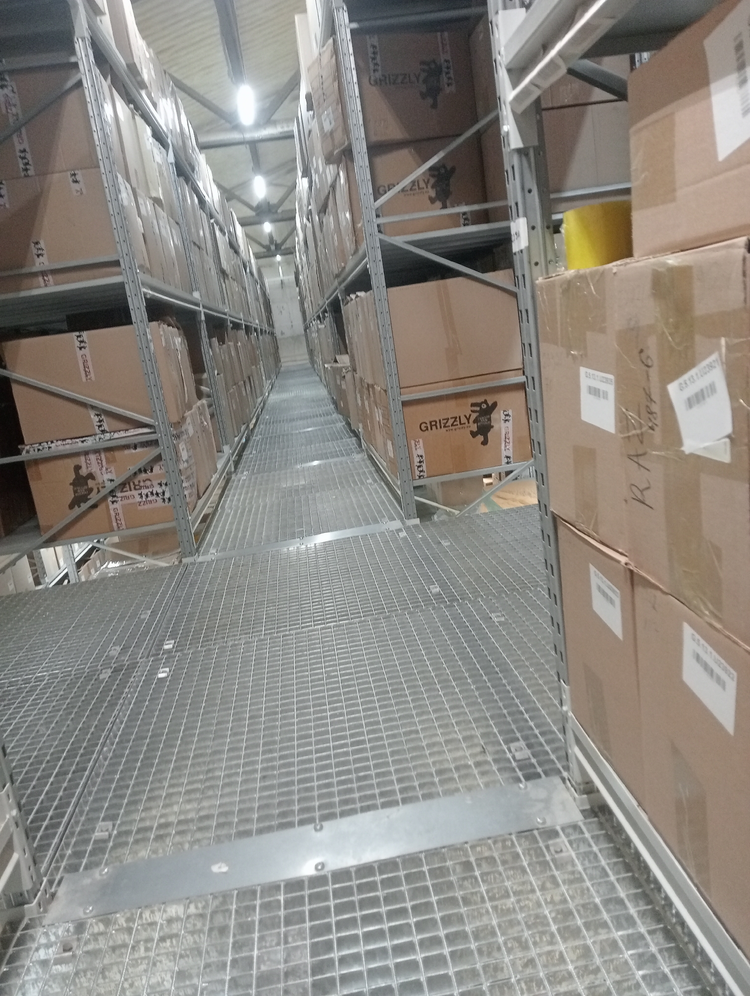 The image shows an interior view of a warehouse with tall shelving units filled with cardboard boxes. The shelves are made of metal, and the flooring appears to be a metal grate. The lighting suggests it is daytime, with artificial lights visible on the ceiling. The boxes are stacked high, some with visible labels that include the brand name "GRIZZLY" and website "www.grizzly.eu". The overall scene depicts a storage facility, likely for manufactured goods. There are no people visible in the image. The location is identified as Aktobe, Kazakhstan.