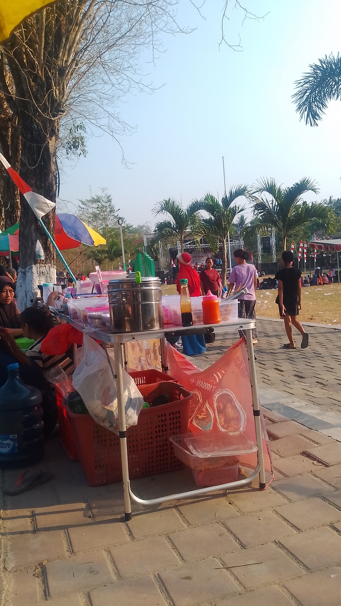 This image captures an outdoor scene in Karanglangu, Indonesia, likely during a daytime event or gathering. In the foreground, a food cart is set up on a brick-paved walkway. The cart holds several items, including large metal containers, plastic containers with food, bottles of sauce, and a red plastic basket filled with various goods. A blue water jug is positioned to the left of the cart. A person wearing a dark outfit is walking away from the cart towards the right side of the image. In the midground, several people are visible, some interacting around other stalls or gathering areas. Behind them, palm trees and other foliage are present, and in the distance, a stage with audio equipment and various banners suggest an ongoing event. The sky is clear and bright. The overall impression is one of a lively outdoor market or festival.