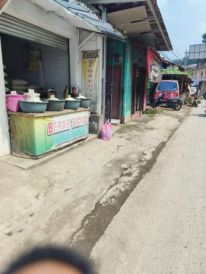 The image shows a small shop on a street in Padalarang, Indonesia (latitude: -6.851, longitude: 107.477). The shop is selling rice, indicated by the large sign "BERAS TIGA PUTIH" and sacks of rice visible in the shop. A woman is sitting inside the shop, likely attending to customers. Four blue plastic basins are arranged in a row on a wooden counter. In the foreground, a pink plastic bag lies on the sidewalk in front of the shop. A red pickup truck is parked on the street in the background, with a motorcycle parked next to it. The sidewalk is made of concrete with a visible crack running down the middle. The street is paved with asphalt. The surrounding buildings are residential and appear to be low-rise with simple constructions. The sky is clear and blue. The image is captured from a low angle, potentially from a person standing on the sidewalk near the shop.