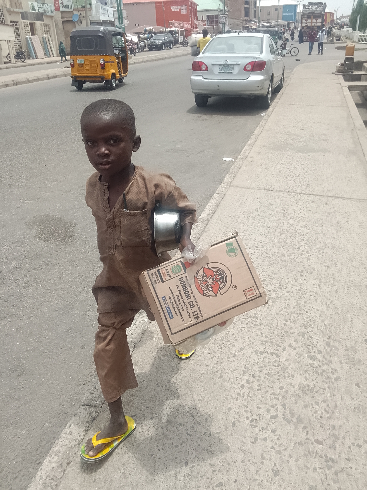 A young boy in Pantame, Nigeria, is seen standing on a sidewalk. He wears a brown, traditional tunic and yellow and black flip-flops. In his left hand, he holds a metal bowl, and in his right, a cardboard box labeled "GONGONI CO. LTD.". The street in the background features cars, a yellow tricycle taxi, and pedestrians. Buildings and shops line the street. The sky is overcast.