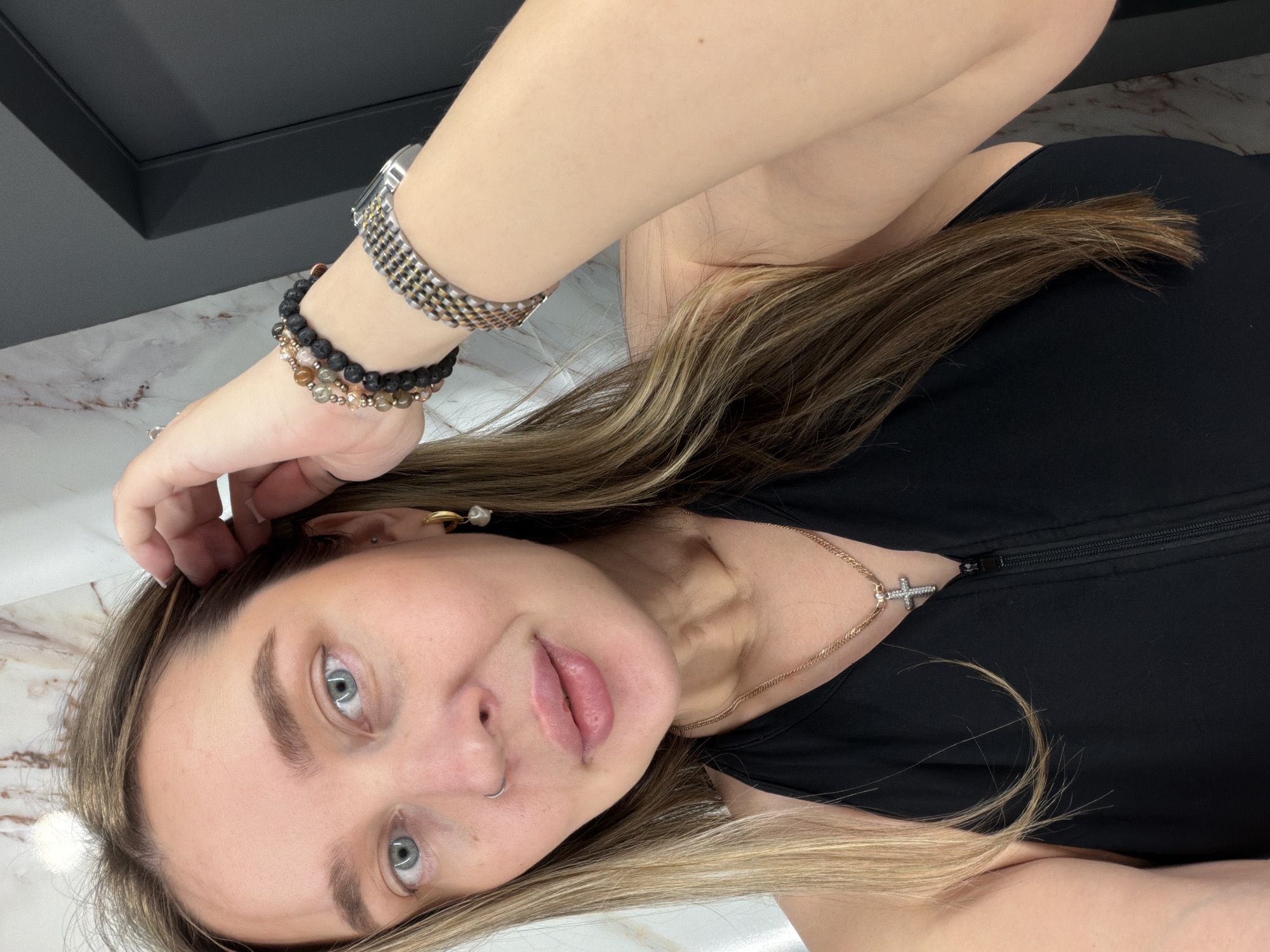 A light-skinned female with blonde highlighted hair, wearing a black top, is positioned at an angle in the frame. She has grey-blue eyes, a nose ring, and a gold cross necklace. Her left arm is raised, displaying a watch and two bracelets on her wrist. The background includes a black element in the upper left corner and a white marble-like surface. The image does not provide information that indicates it was taken in Moscow, Russia.