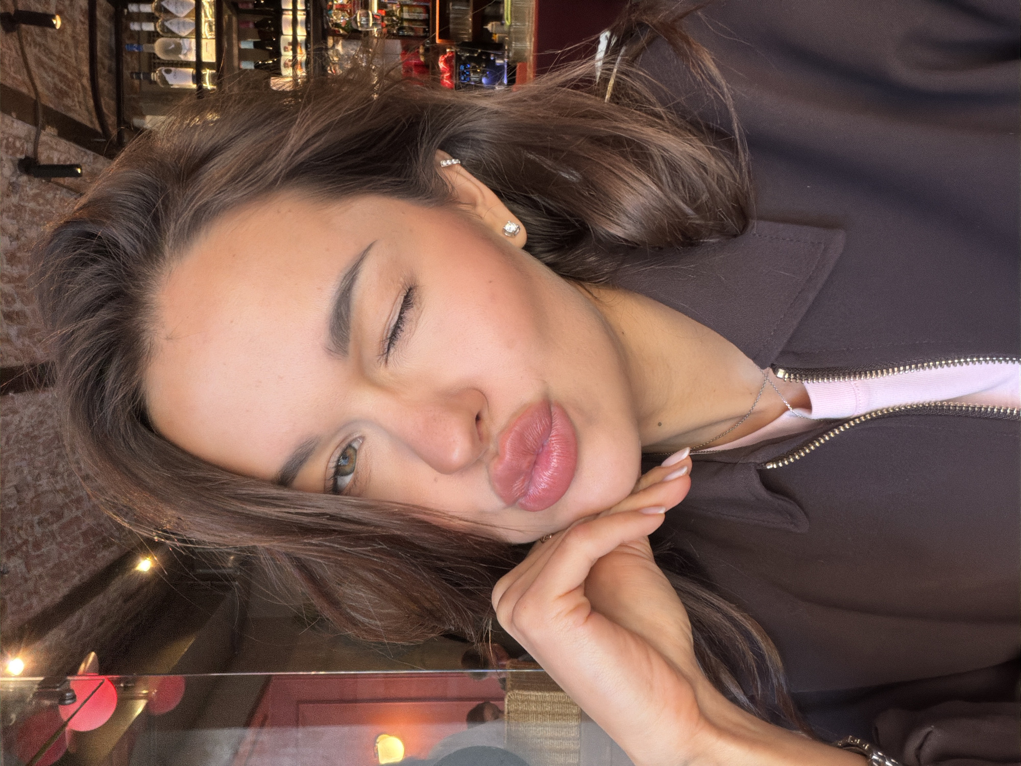 A young woman is taking a selfie in a dimly lit establishment with exposed brick walls and shelves stocked with bottles. She is wearing a dark brown jacket over a pink shirt and has her hand resting on her chin. Her lips are pursed, and she is winking. The lighting suggests it is nighttime, and the background indicates a bar or restaurant setting. The specific location within Saint Petersburg is not determinable from the image.