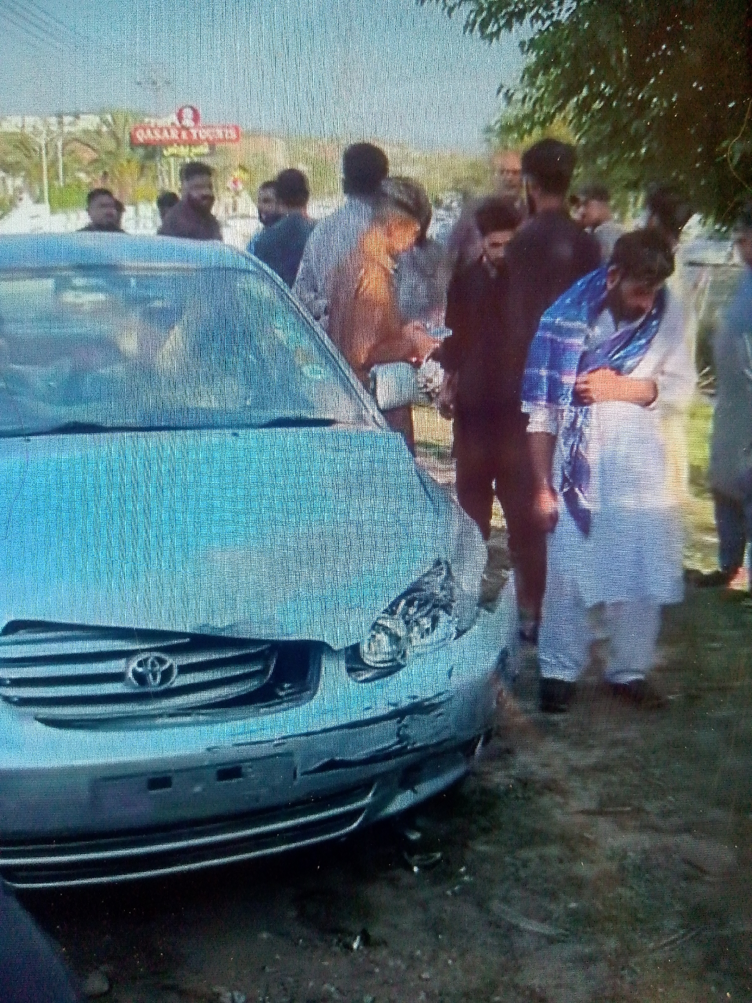 A group of approximately eight to ten men are gathered around a silver-blue Toyota sedan, which appears to be damaged at the front. The car, likely a Corolla model given the visible emblem and grille, has a dislodged or broken front bumper and damage around its passenger-side headlight. The men are standing on a dirt or gravel surface next to the car, with some looking towards the vehicle. One man in the foreground on the right is wearing a white shalwar kameez and a blue and white patterned shawl draped over his shoulders. Other individuals are dressed in dark or earth-toned clothing.
The scene is outdoors, brightly lit by natural daylight, suggesting a clear day. In the background, trees and foliage are visible, along with a structure that features a prominent red sign. On this sign, the words "QASAR & TOURIST" are partially discernible, with some additional text in what appears to be Urdu or Arabic script below it, though it is largely unreadable due to pixelation. The overall impression is of people gathered to observe or discuss the damaged vehicle.
