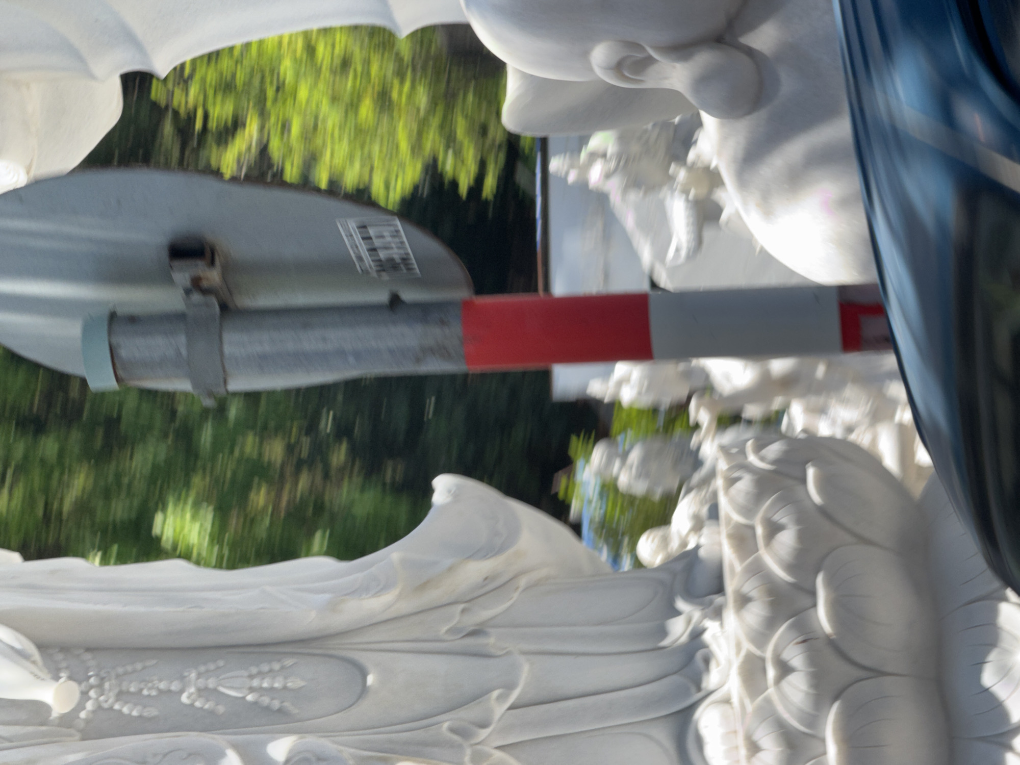 A blurred, close-up photograph shows a white marble statue. The statue's features, including part of a face and draped cloth, are visible. Behind the statue, partially obscuring it, is a signpost. The signpost has a circular gray sign and a gray and red striped pole. In the background, blurred green foliage suggests trees or bushes. The image is sideways, with the top of the statue pointing to the right. It is likely that this image was taken at the Lady Buddha (Quan Am) statue located at the Linh Ung Pagoda in Da Nang, Vietnam.
