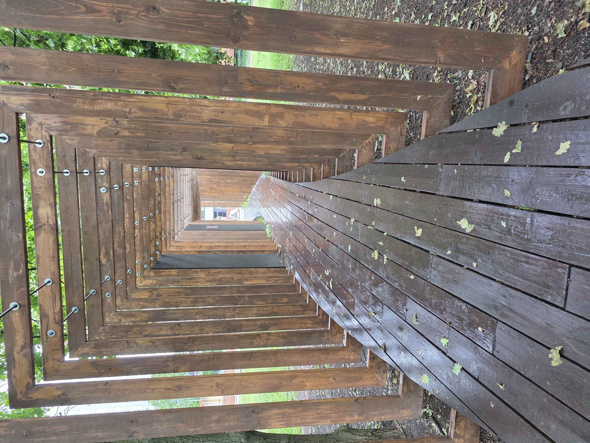An outdoor scene features a long, angular wooden structure resembling a tunnel or pathway, viewed from one end looking towards the other. The structure is made of numerous horizontal wooden planks, forming repeating square or rectangular frames that progressively narrow into the distance, creating a strong sense of perspective.
The sides of the pathway are slatted, allowing views of vibrant green foliage and tree branches through the gaps. On what appears to be the upper-left side of the tunnel (due to the image's rotation), metal rods secured with circular washers are visible, extending horizontally between the wooden frames, likely serving as structural supports or design features. The floor of the pathway is made of dark wooden planks, which appear wet and reflective, indicating recent rain or damp conditions. Light green leaves are scattered sparsely on the wet wooden floor and on the dark, natural ground surrounding the structure, which is covered with dirt and more fallen leaves.
In the far distance, at the end of the narrowing wooden frames, a faint glimpse of a light-colored building with a dark window can be seen. The scene is illuminated by natural daylight. No people are present, and no specific activity is taking place. There is no visible text.