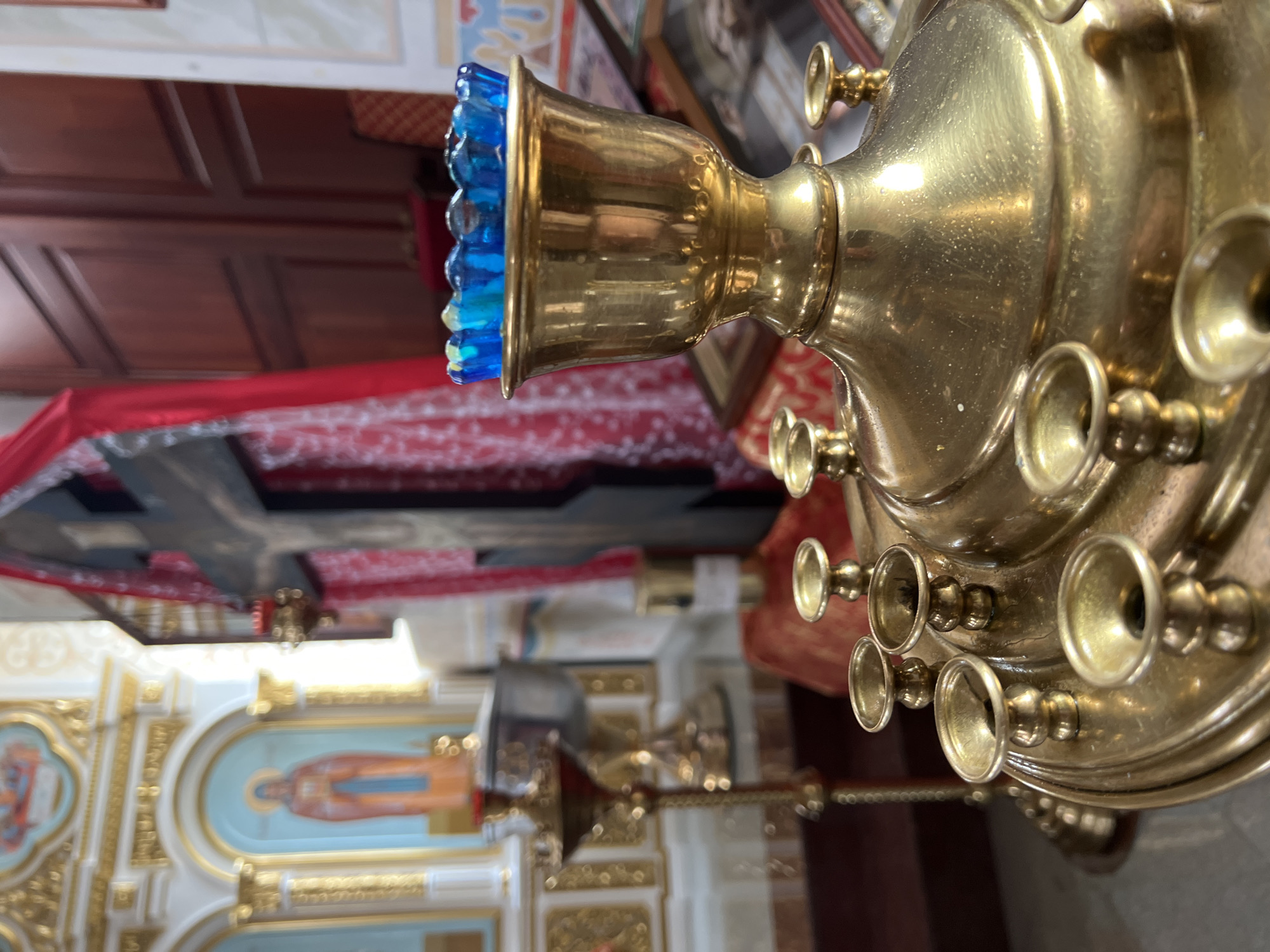 The image shows an interior scene of a Russian Orthodox church, potentially located in Kargopolye, Russia. The foreground is dominated by a brass candle chandelier with multiple candle holders and a blue glass rim. In the background, there's a red cloth-covered cross, a mural of a saint, and other religious adornments typical of Orthodox churches. The lighting is soft and somewhat diffused, suggesting natural light filtering through the church's windows.