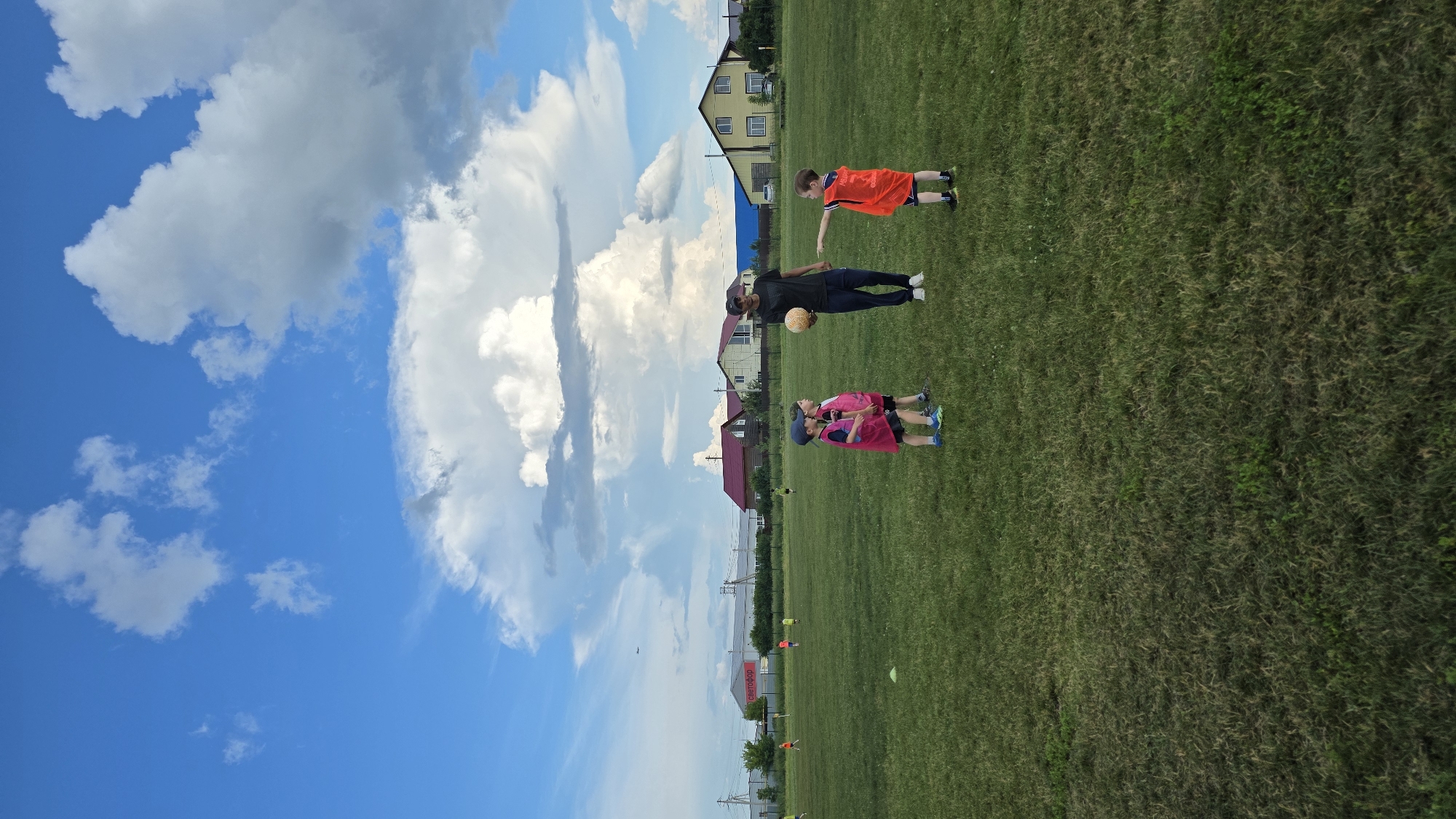In Bulgakovo, Russia, a man and three young children are on a grassy sports field under a cloudy blue sky. The man, wearing a dark t-shirt and pants, holds a light-colored soccer ball. Two children in pink vests and blue caps stand to his left, and one child in an orange vest stands to his right, reaching out. The field has green grass with scattered patches of dry grass and some white cones. In the background, several houses with red and yellow roofs are visible, along with a large building displaying a sign with Russian text that reads "СВЕТОФОР". A small airplane is visible in the distance in the sky.