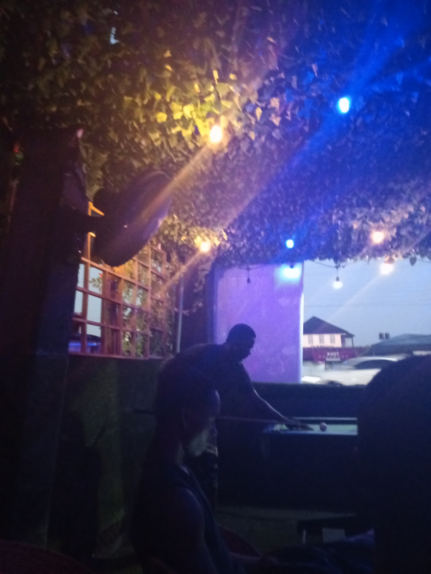The image depicts an outdoor pool hall in Ado, Nigeria, at dusk. The space is dimly lit with a mix of blue and yellow string lights strung through a leafy covering overhead. Two men are visible: one is playing pool at a table, and the other is seated in the foreground. Another person is partially visible in the lower right corner of the frame. A fan is mounted on a wooden lattice structure, suggesting this is an outdoor space. In the background, outside the covered area, a building and cars are visible, indicating a cityscape.
