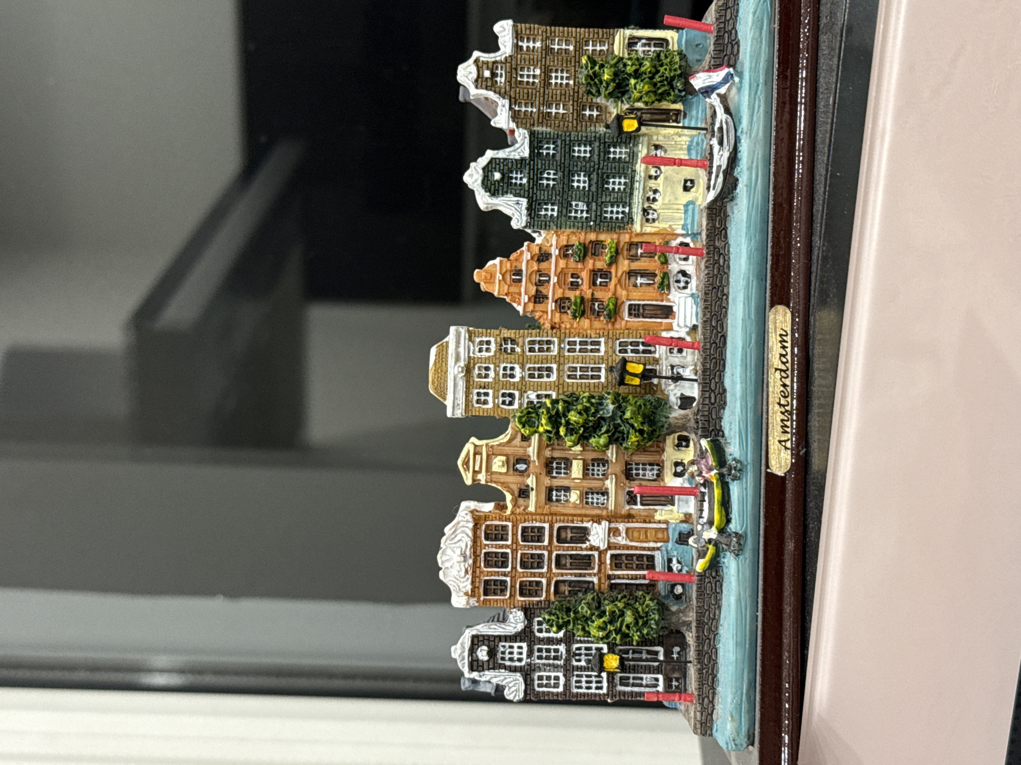 This is a close-up, eye-level, medium shot of a miniature model of Amsterdam houses, set on a wooden base. The model depicts a row of narrow, tall, Dutch-style buildings with colorful facades, intricate details like windows, doors, and decorative elements. The buildings are situated along a canal with small boats, including a yellow rowboat with two figures and a white boat with a Dutch flag. Lush green trees are interspersed among the buildings, and red lampposts line the canal. The base of the model has a gold plaque with the word "Amsterdam" inscribed on it. The background is blurred, showing a dark, reflective surface that suggests an indoor setting, possibly a window or a screen. There is no indication of Volgograd, Russia in the image.