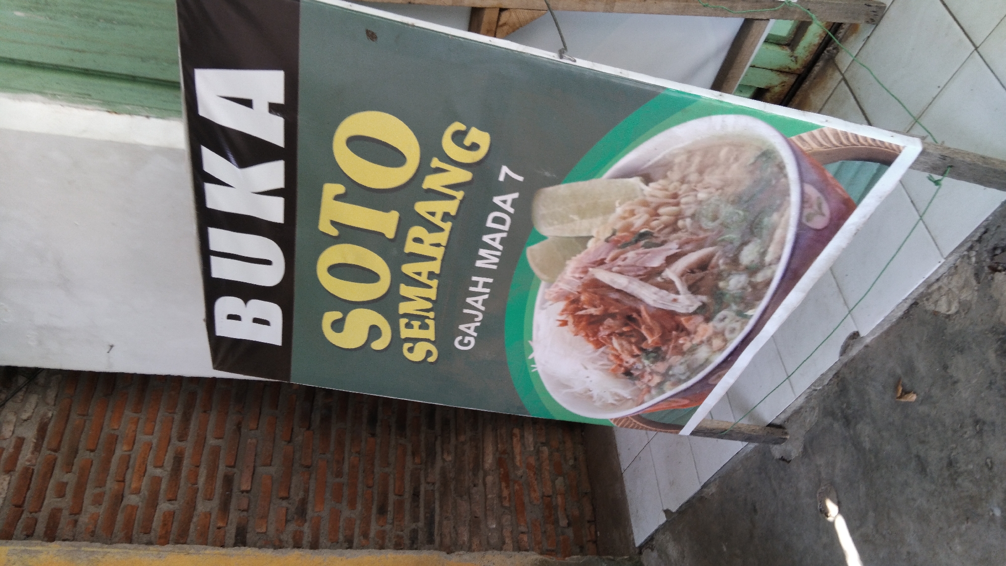 A weathered, leaning A-frame sign advertises "BUKA SOTO SEMARANG GAJAH MADA 7." The sign features a large, appetizing image of a bowl of soto, a traditional Indonesian soup, filled with shredded meat, noodles, and broth, topped with two pieces of what appear to be boiled eggs or tofu. The sign is partially obscured by a weathered brick wall on the left and a peeling green door or window frame on the right. The ground is a mixture of cracked concrete and white tiles. The overall impression is one of a humble, local eatery.