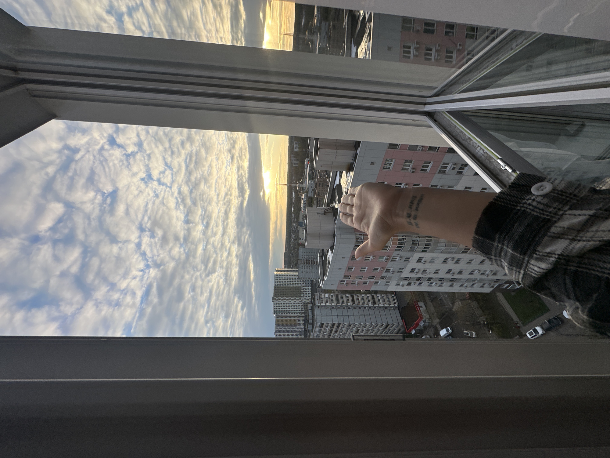 The image, rotated 90 degrees counter-clockwise, captures an urban panorama from a high vantage point, looking out through an open window. In the immediate foreground, a person's right arm and hand are extended, reaching out of the window opening. The person is wearing a plaid shirt. A tattoo is visible on the back of the wrist/hand, showing text fragments such as "04.02.04", "DMY", and "AM", along with numbers that appear to be "13-14" or "13.14".
The sturdy grey window frame defines the bottom and right edges of the view. Beyond the window, a dense cityscape of multi-story apartment buildings dominates the midground. Many buildings are light-colored, some with pinkish hues, and feature numerous windows and air conditioning units. Several cars are parked on the ground level below, next to what appears to be a small green area or park.
The sky in the background is filled with scattered, textured clouds, illuminated by the warm, golden light of what appears to be a sunset or sunrise on the horizon. Reflections of the buildings and sky are visible on the window panes to the upper right. The scene depicts a typical residential area in Moscow, Russia, as suggested by the context.