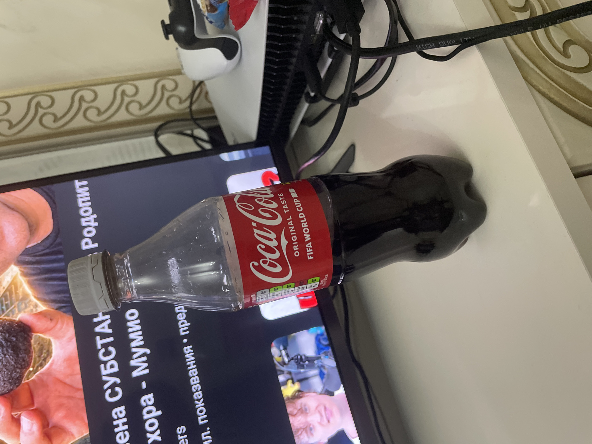 The image, taken in Ihtiman, Bulgaria, captures a casual indoor setting. A plastic Coca-Cola bottle, partially filled with the dark beverage, lies horizontally on a white surface, possibly a shelf or table. The bottle's label is prominent, displaying "Coca-Cola ORIGINAL TASTE FIFA WORLD CUP" in red and white, along with a nutrition information table.
Behind the bottle, a television screen displays content, likely a video, with Cyrillic text. Visible text includes "Родопит", "СУБСТАН" (SUBSTANCE), "хора", "Мумио" (Mumiyo or Shilajit), "ил. показвания" (impressions/views), and "пред". The video shows a person's hand holding a dark, irregular object.
To the upper right, a white electronic device, resembling a PlayStation console, is partially visible, surrounded by tangled black cables. A decorative light-colored wall featuring a golden, repeating wave-patterned trim is in the background. A black cable with "HIGH QUALITY" printed on it crosses the upper right portion of the frame. The scene suggests a moment of leisure, possibly involving television viewing or gaming. No specific time of day or emotions are discernible.