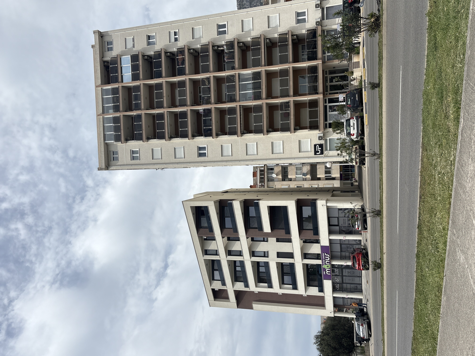 The image shows two buildings in Bar, Montenegro, standing alongside a road and sidewalk. The building closer to the viewer is a modern, multi-story structure with brown and white facade featuring projecting rectangular balconies with dark glass. A purple sign for "Manus" is visible on one side. A red car is parked in front of the building. Further along the road, a taller, older building with a concrete facade and recessed balconies stands, with satellite dishes visible on its roof. Cars are parked outside the base of this building. The sky is overcast with clouds. The photograph appears to have been taken from a slightly elevated position across the road from the buildings.