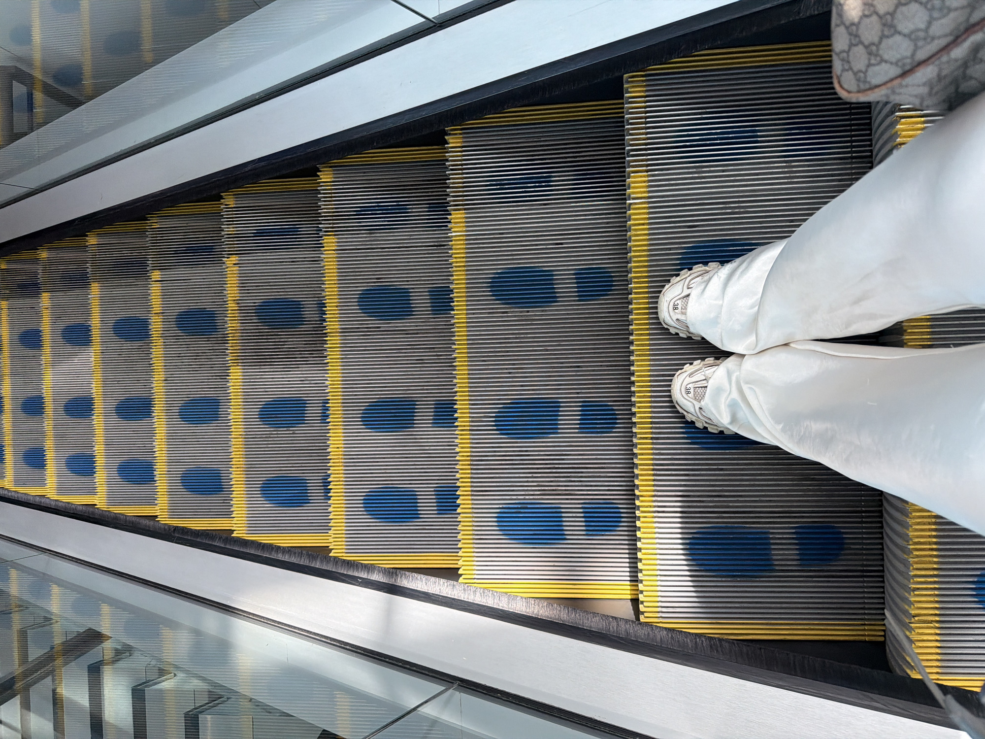 The image shows a top-down view of an escalator with a person standing on it. The escalator steps are gray with yellow edges and blue footprints painted on them. The person is wearing white pants and white sneakers. A patterned bag is partially visible on the top right of the image. The escalator has clear glass sides. The location is not specified as Uttlesford, United Kingdom.