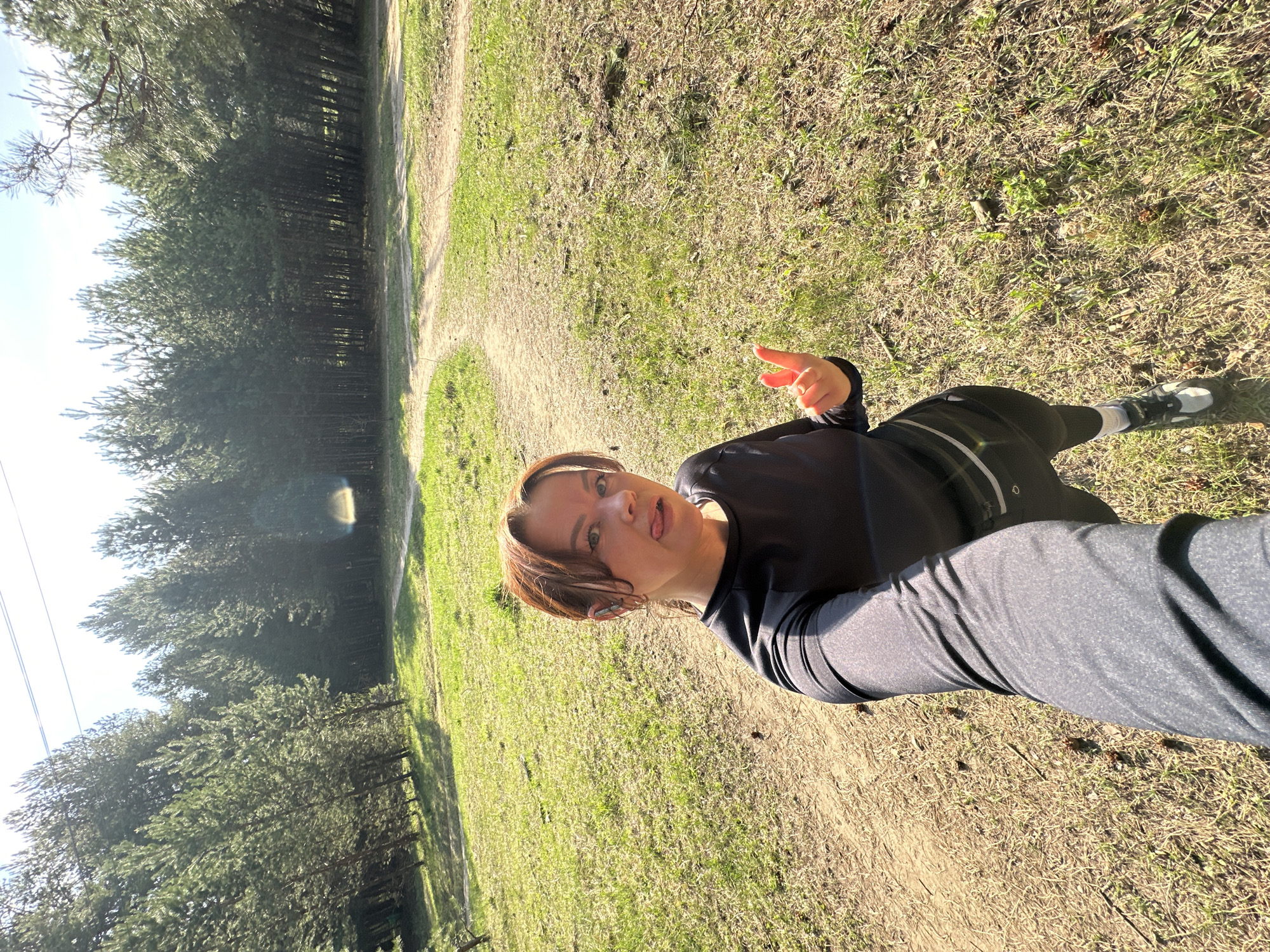 The image shows a person standing on a grassy, dirt path in a wooded area. The person is wearing dark athletic clothing and makes a peace sign with their hand. The background consists of tall trees on the left and open grassy areas on the right. It is daytime with bright sunlight. The location is likely within a natural area in Chaikovsky, Russia.