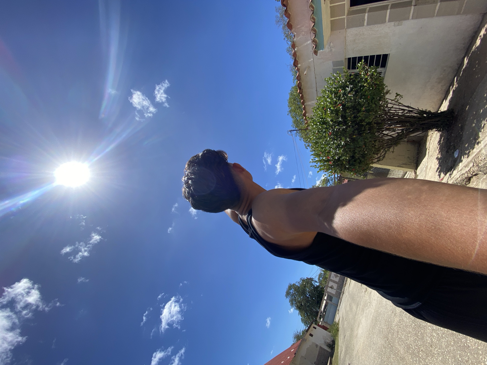 A low-angle, brightly lit outdoor shot shows a person looking up at a blue sky with scattered clouds and the sun. The person has dark hair, dark skin, and wears a black tank top. The sky is a clear blue color with some small, puffy white clouds. The sun is shining brightly in the upper left corner of the frame, creating lens flare. Buildings and greenery are visible in the background, including a house with a red tile roof and a small tree. The location appears to be a sunny area in Juan Griego, Venezuela.