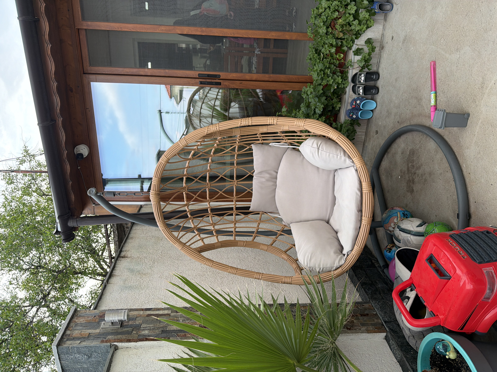 The image shows a patio or backyard area adjacent to a building, potentially located in Burgas, Bulgaria, given the request. A wicker egg chair with light gray cushions sits on the paved ground. The chair is positioned near a wall with stone accents and some plants. There's a red plastic toy truck and several balls stored behind the chair base. Children's footwear are on the ground near the building. A gray tubing object sits next to the chair. The building has a wooden framed sliding glass door. A security camera is visible near the top of the wall. A tree is visible on the left of the image.