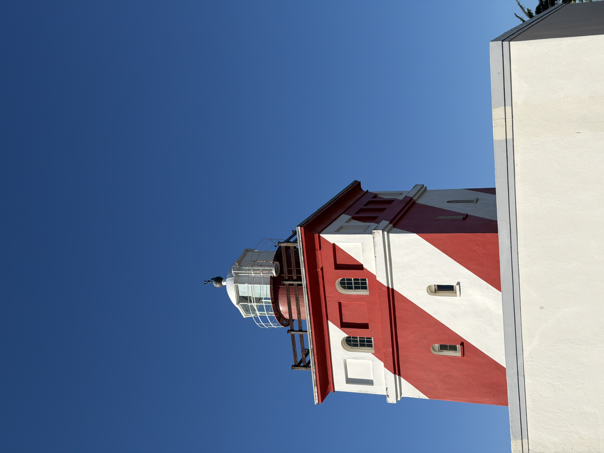 The image shows the red and white striped Milnerton Lighthouse in Cape Town, South Africa, against a clear blue sky. Part of an adjacent white building is visible on the right.