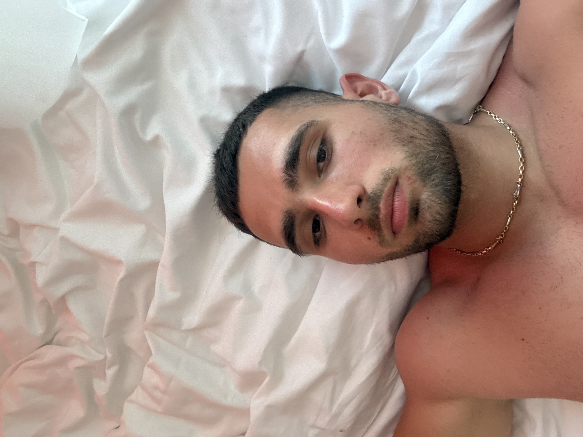 This is a close-up, overhead shot of a young man lying on a white bed. He has short dark hair, a beard, and is wearing a gold chain. The bedsheets are rumpled, suggesting a relaxed or recently occupied state. The lighting is soft and diffused, hinting at an indoor setting, possibly a bedroom. The man's expression is neutral, and his eyes are partially closed. The overall mood is intimate and serene. There is no visible text in the image.